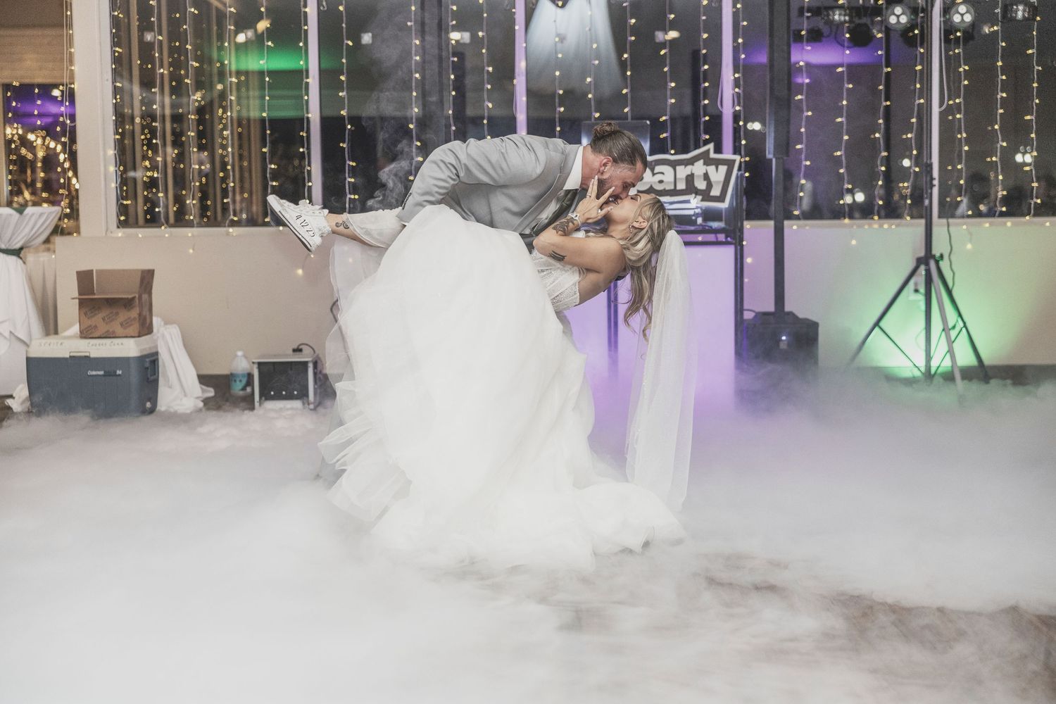 Couple shares romantic first dance on misty dance floor with purple and white lighting at wedding reception.