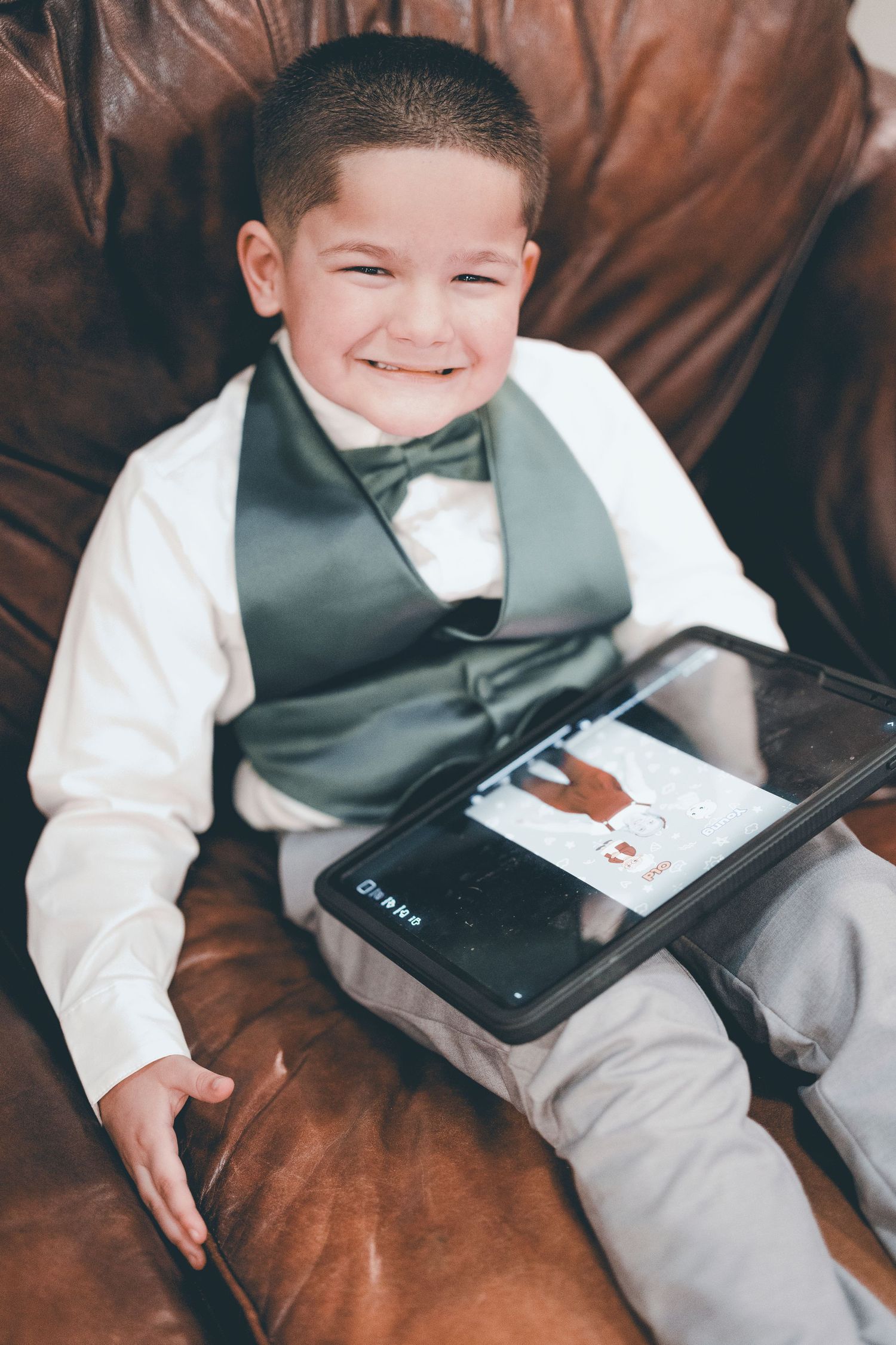 Young child in formal wear with bowtie lounges on brown leather couch holding a tablet device while smiling.