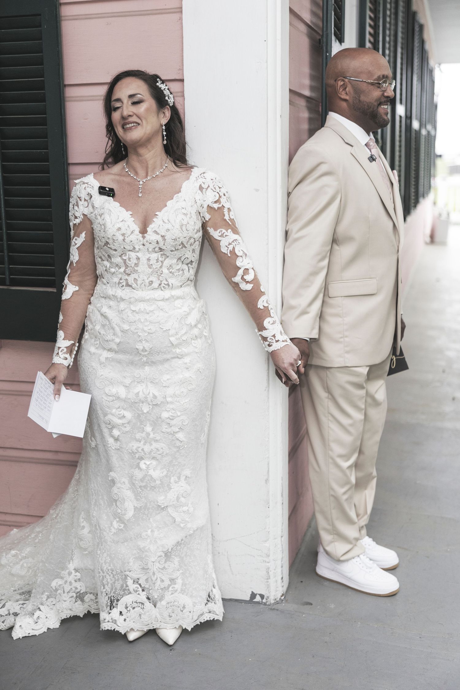 A bride in lace long sleeve wedding gown and groom in tan suit hold hands while standing on opposite sides of a pink wall.