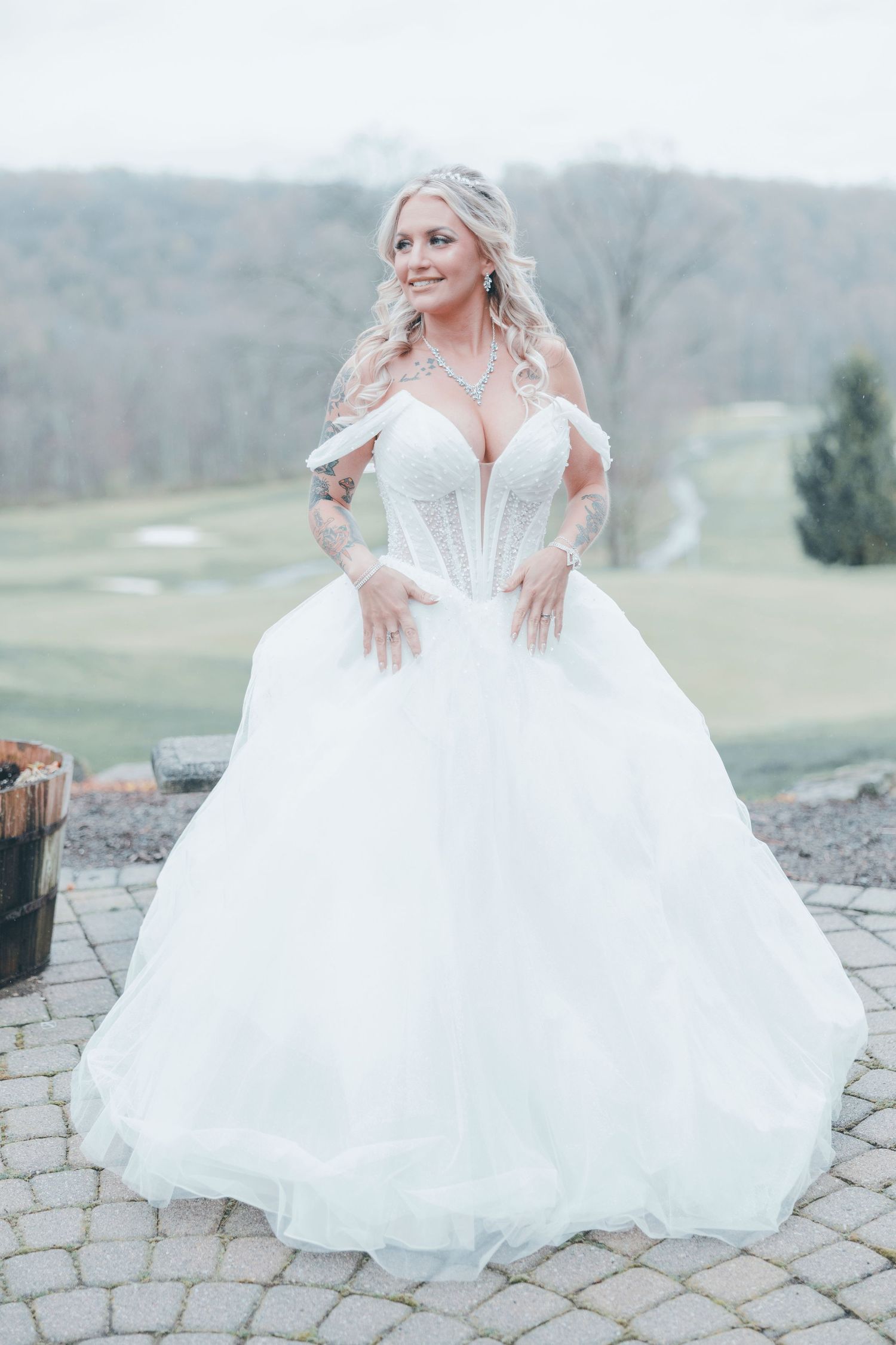 A bride in a luxurious white off-shoulder ballgown wedding dress poses on stone pavers against a misty outdoor backdrop.