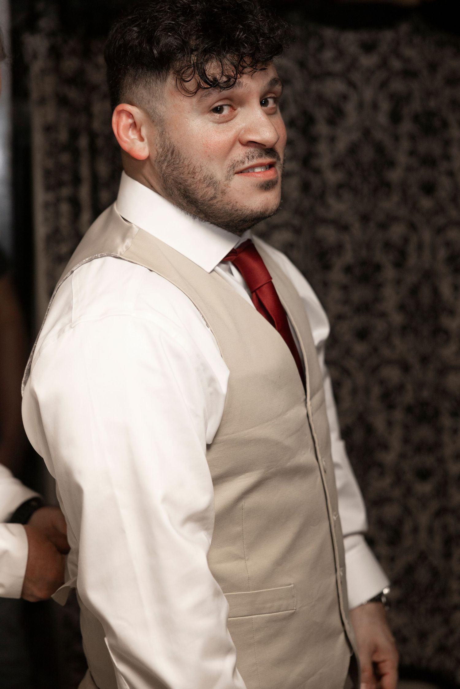 A well-dressed person in a beige waistcoat and red tie smiles while looking off to the side.
