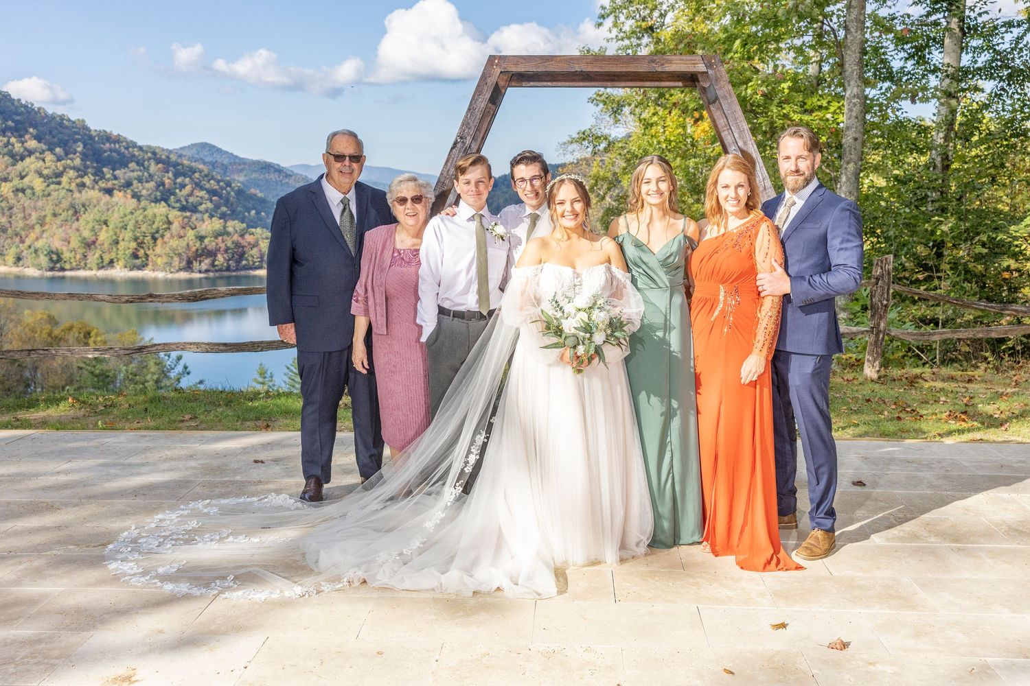 A wedding party poses together on a mountain overlook with a scenic lake and forest backdrop on a sunny day.