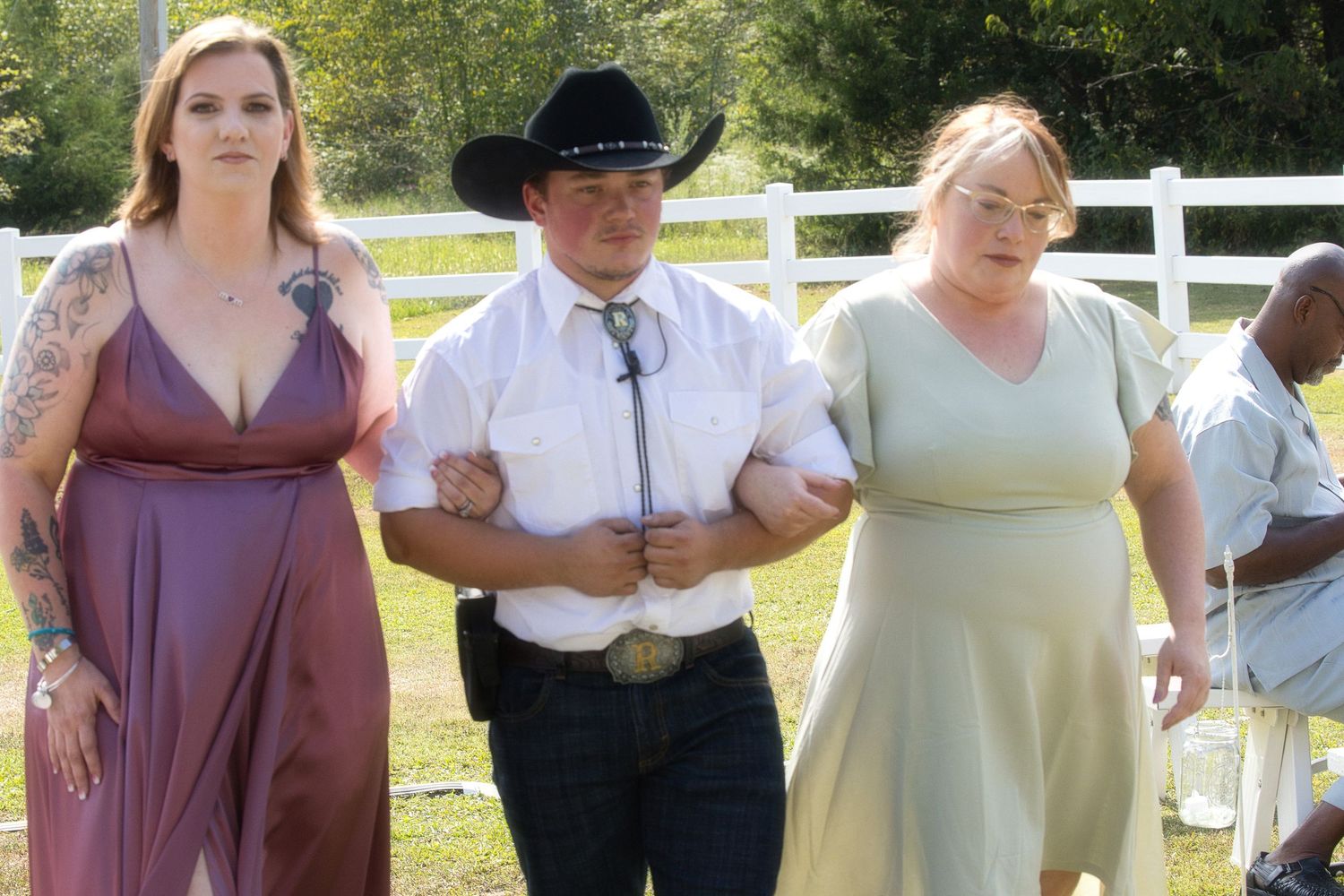 Wedding guests in formal attire walking arm in arm together along a white fence outdoors.