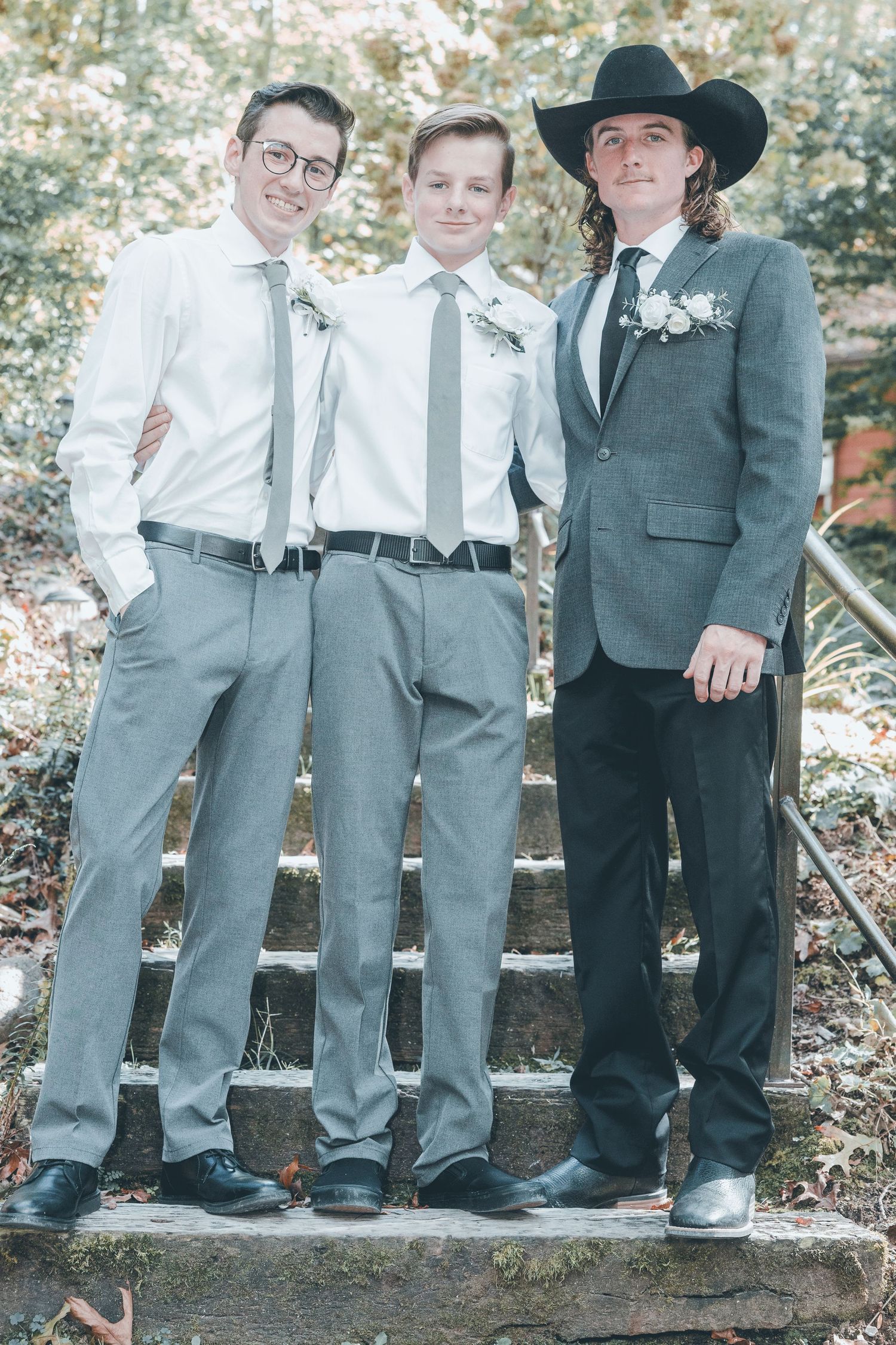 Three people stand together at a wedding wearing coordinated gray suits and ties, with one person in a black cowboy hat.