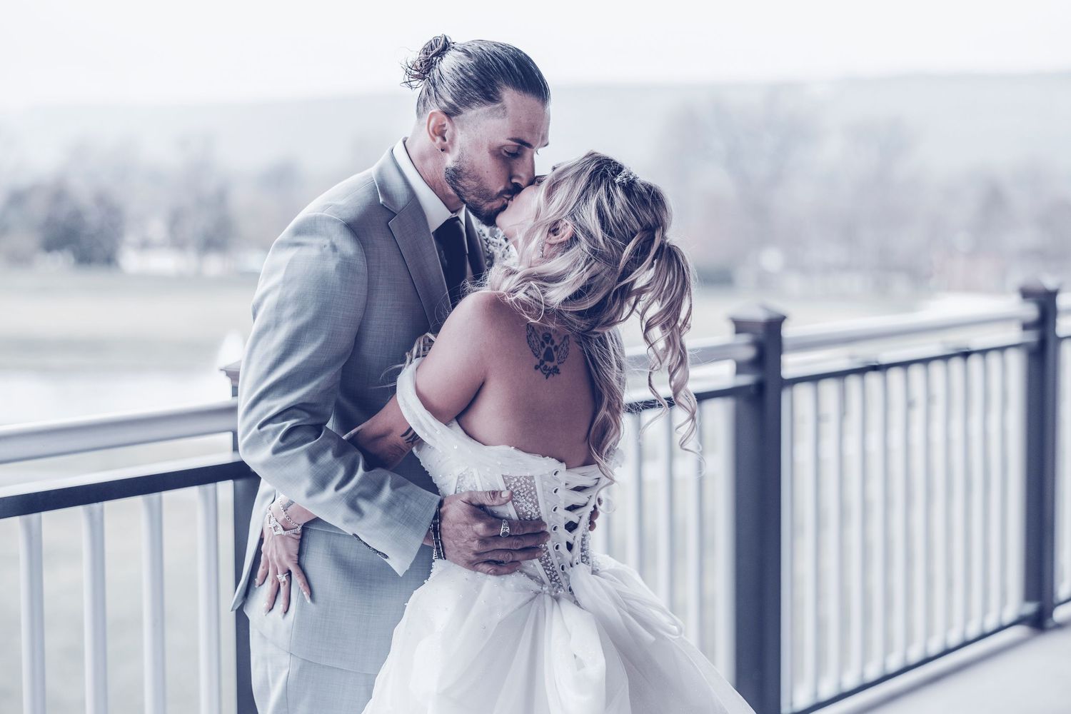 A romantic wedding couple embraces on a bridge railing during a misty winter day.