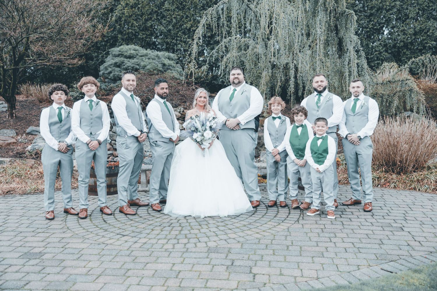 Wedding party in coordinated gray and white attire pose together on stone pathway beneath weeping willow tree.
