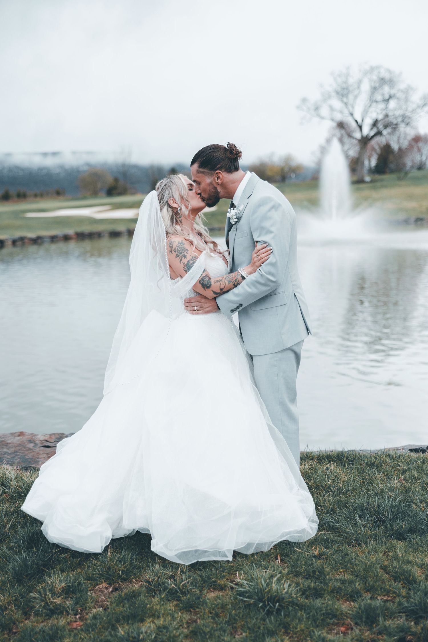 A romantic wedding photo of a couple embracing by a lake with a fountain in the background.