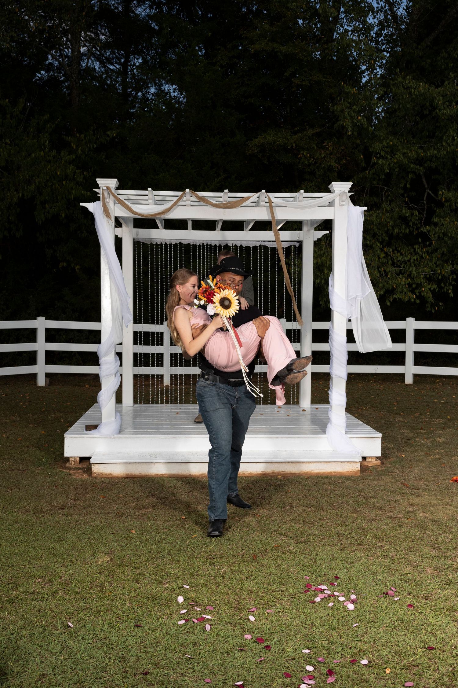 A romantic moment under a white pergola with sunflowers and scattered rose petals on the grass.