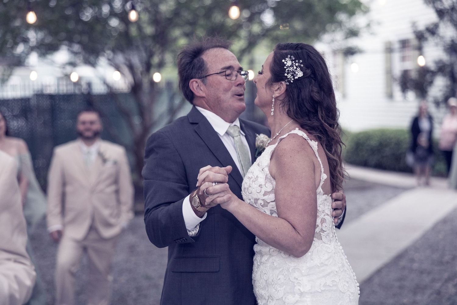 A joyful couple dances together on their wedding day, the bride in white lace and groom in a dark suit outdoors.