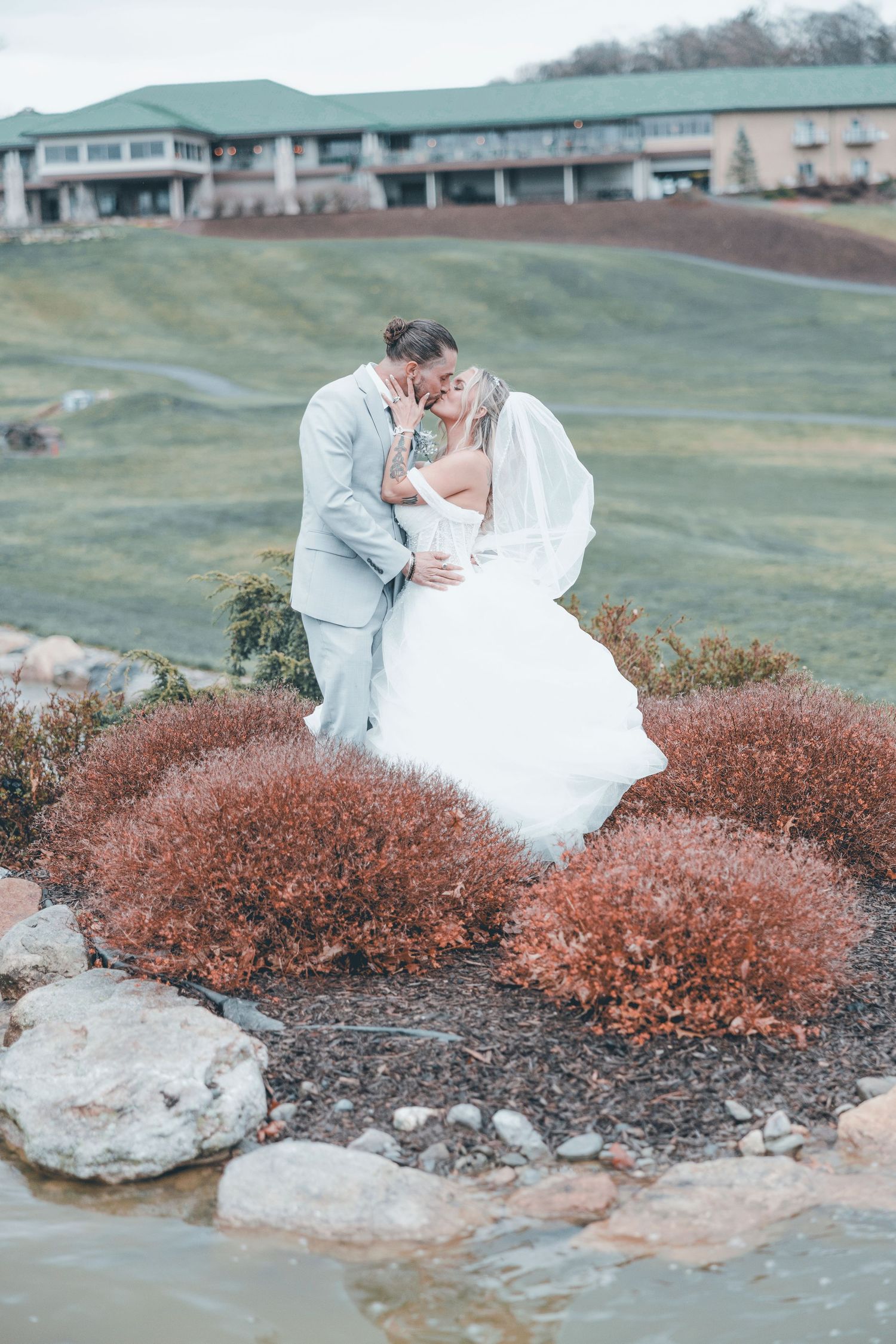 Newlyweds share tender moment among autumn-colored landscaping and rocks near a golf course clubhouse.