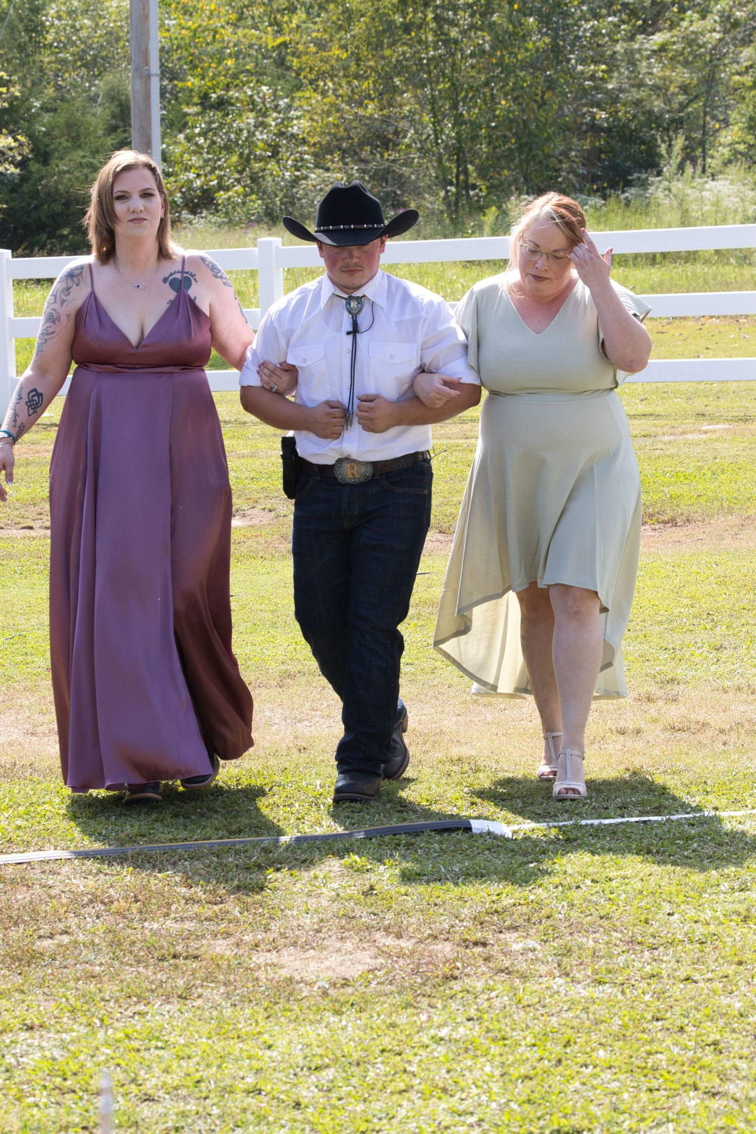 Three formal wedding guests in mauve, black, and mint-colored attire walk together along a sunny fence-lined path outdoors.
