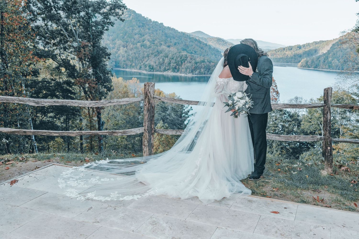 A romantic wedding moment overlooking a serene mountain lake with a flowing white gown and rustic wooden fence in view.