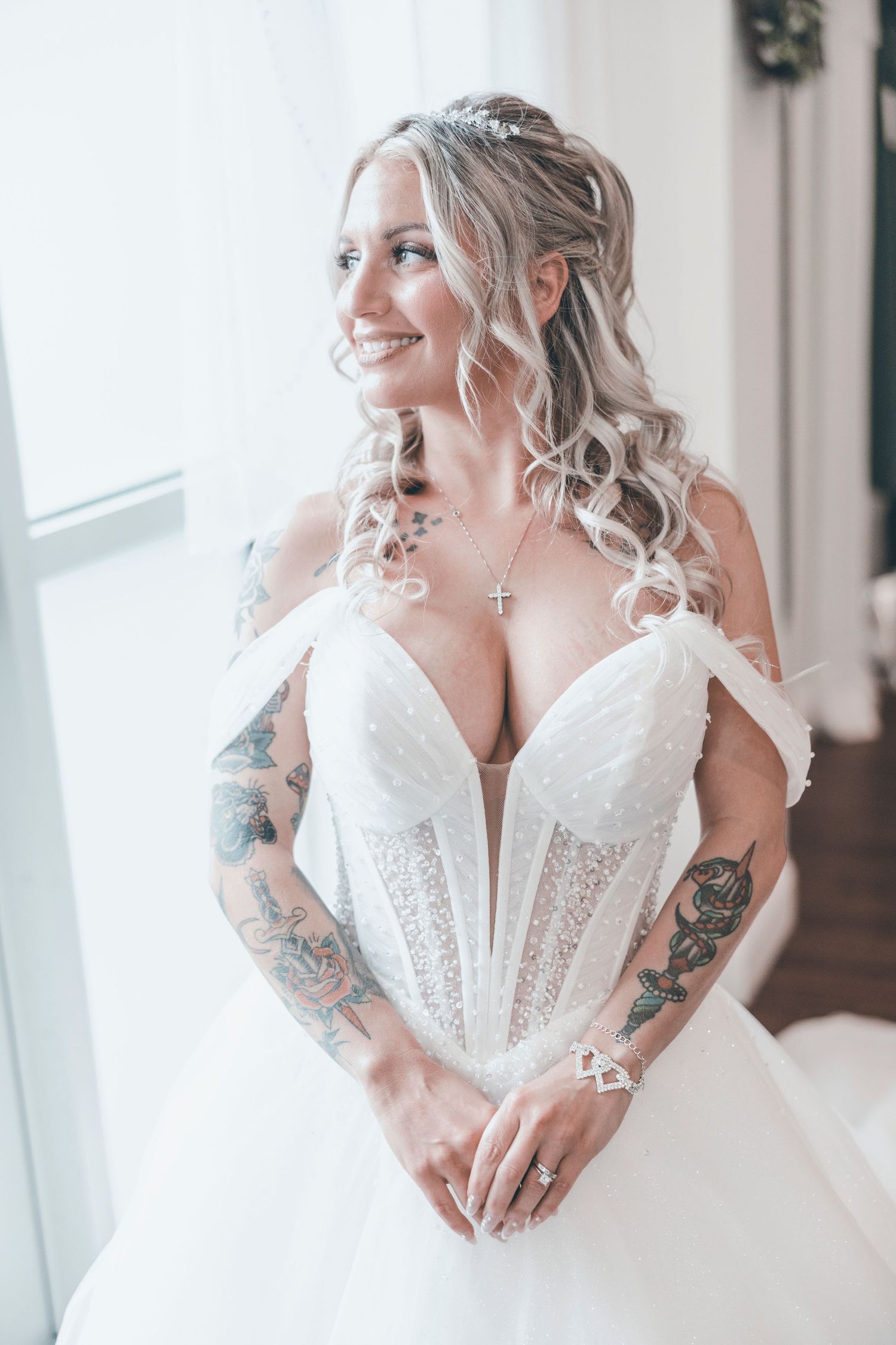 Beautiful bride in white corset wedding dress with lace details and tattoo sleeves poses by window in soft light.