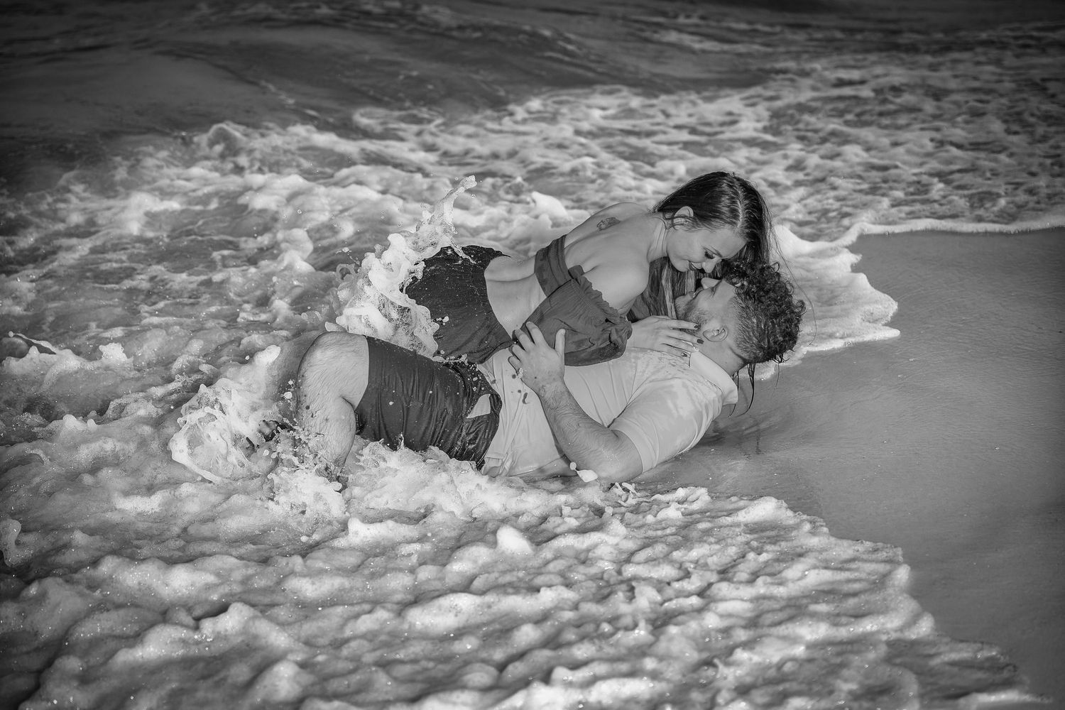 Romantic couple lying in the surf as ocean waves wash over them on a beach in black and white photography.