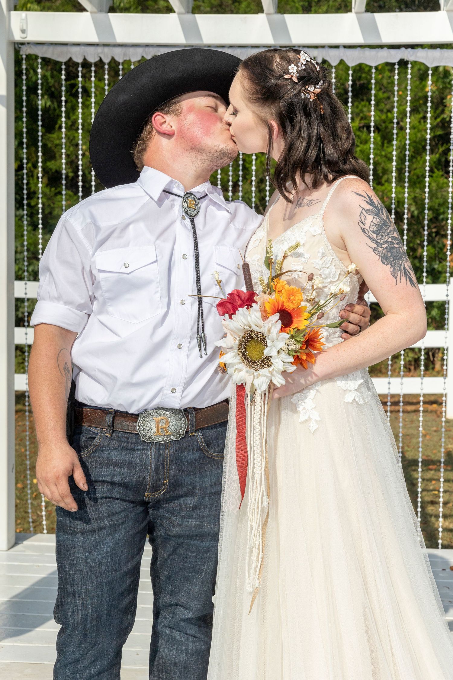 A couple shares a romantic kiss under a white pergola decorated with flowers at their country wedding.