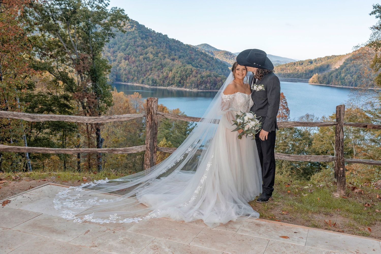 A couple shares a romantic moment near a scenic lake overlook with mountains in the background during autumn.