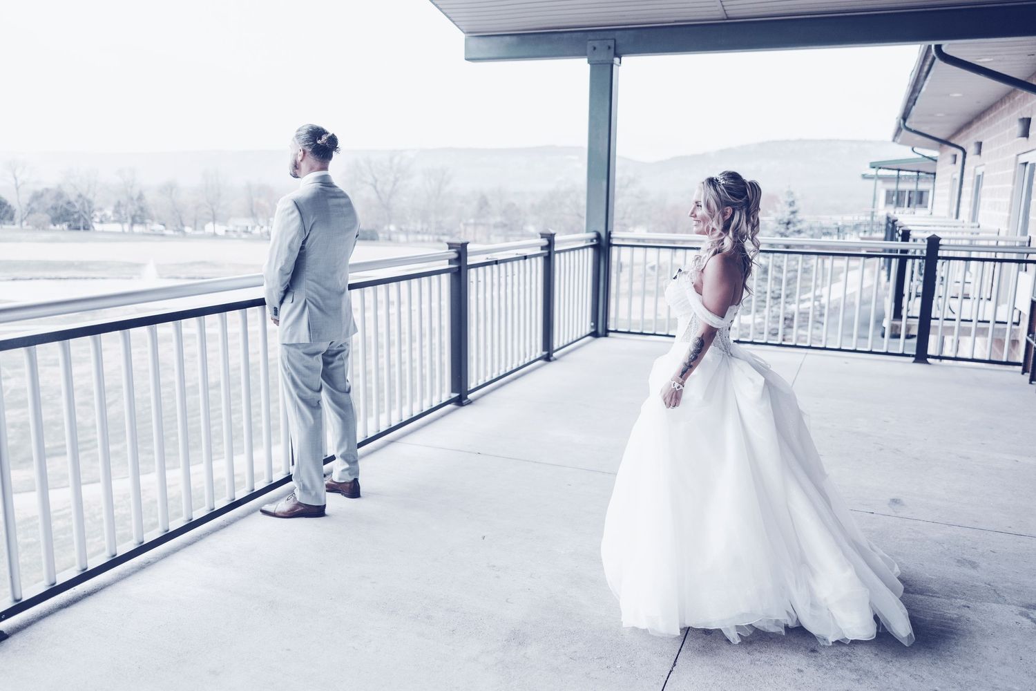 A bride in a flowing white gown stands on a covered balcony while her groom faces away on a cloudy day.