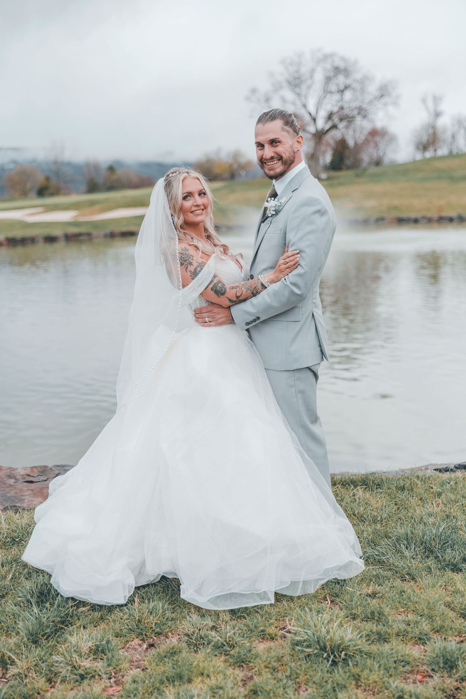 A couple in wedding attire poses by a serene lake on an autumn day with muted natural colors in the background.