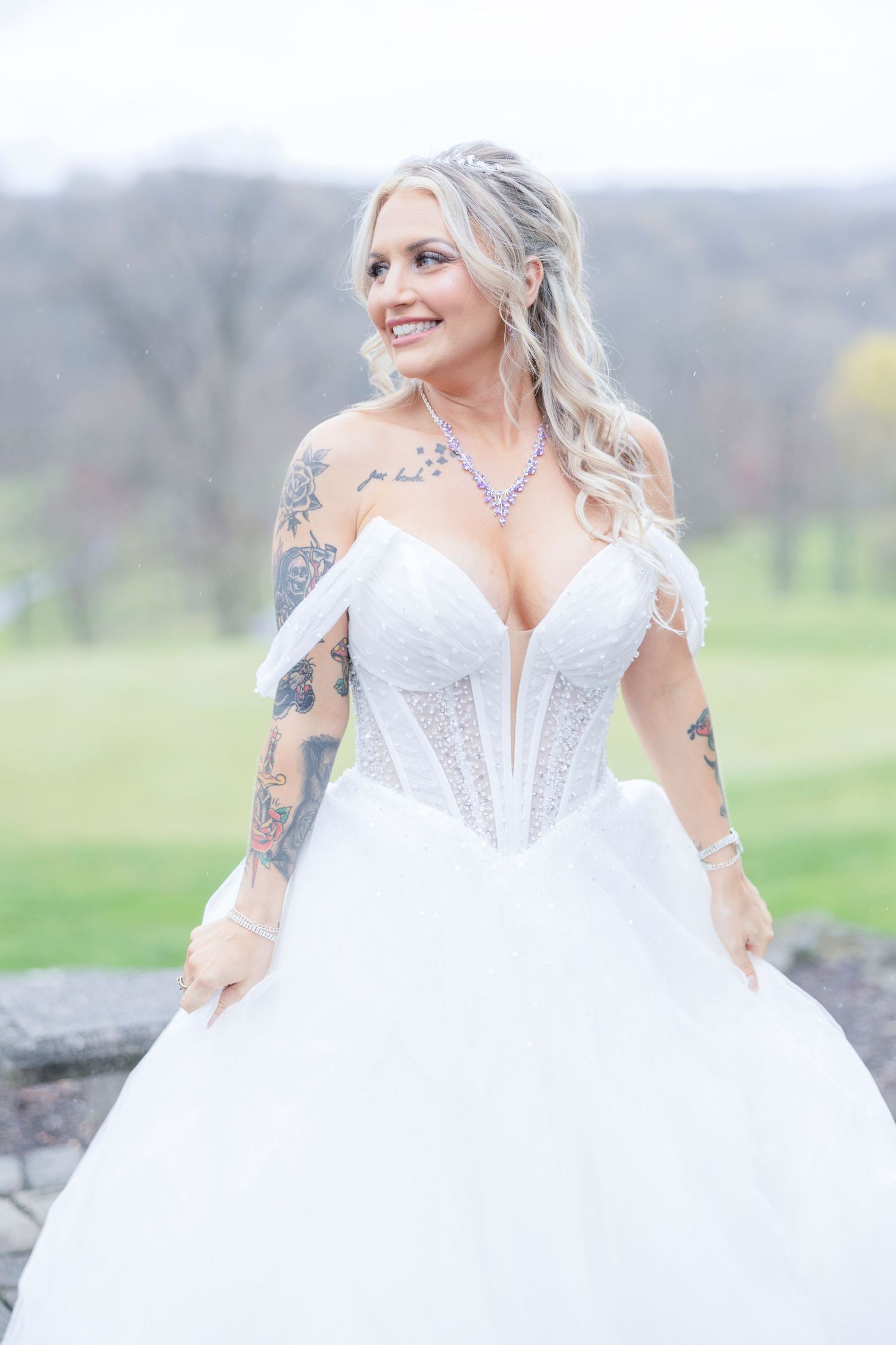 A bride in a white corset ball gown with tattoos poses outdoors in a natural setting.