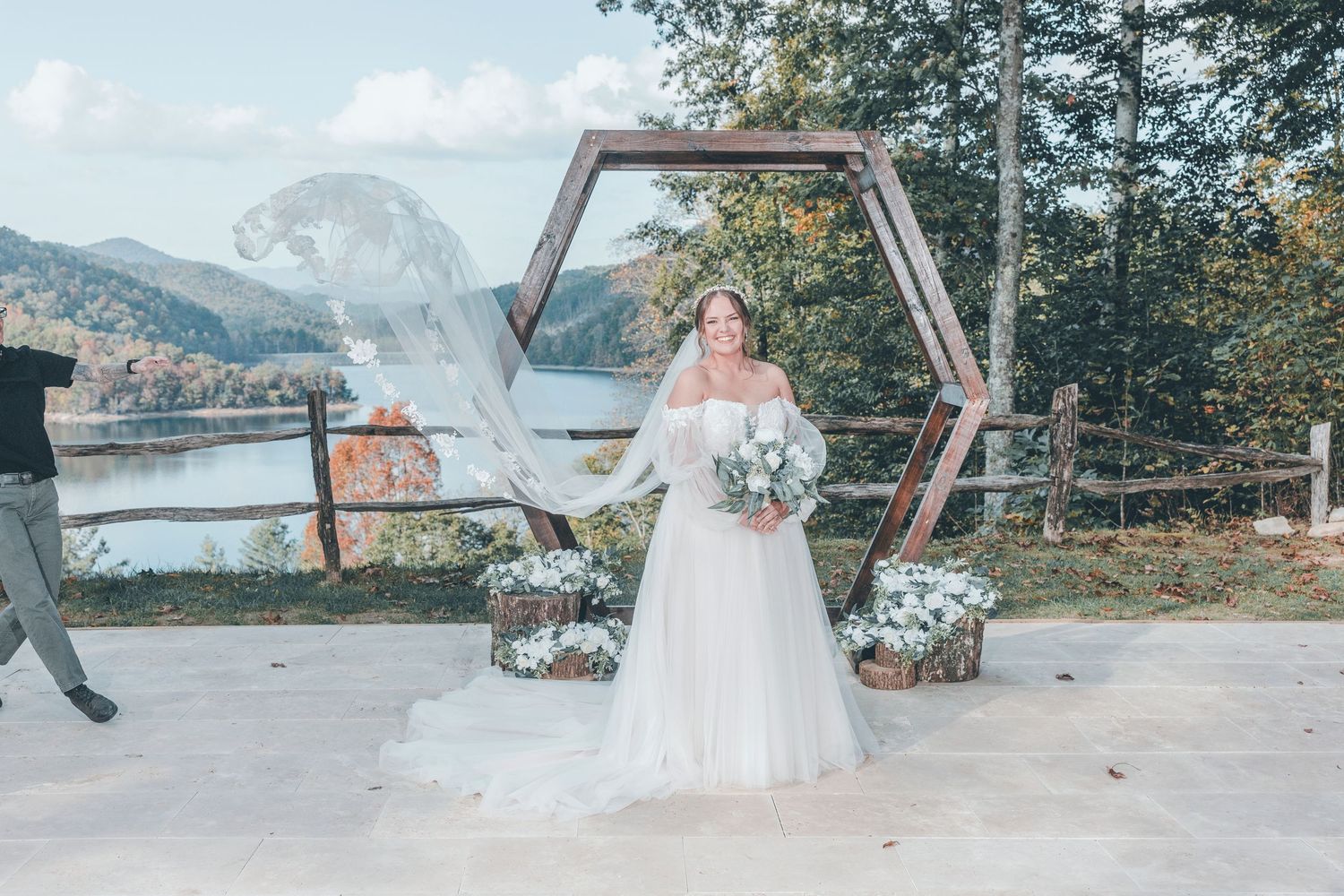 A scenic mountain wedding ceremony featuring a geometric wooden arch decorated with flowers against a lake backdrop.