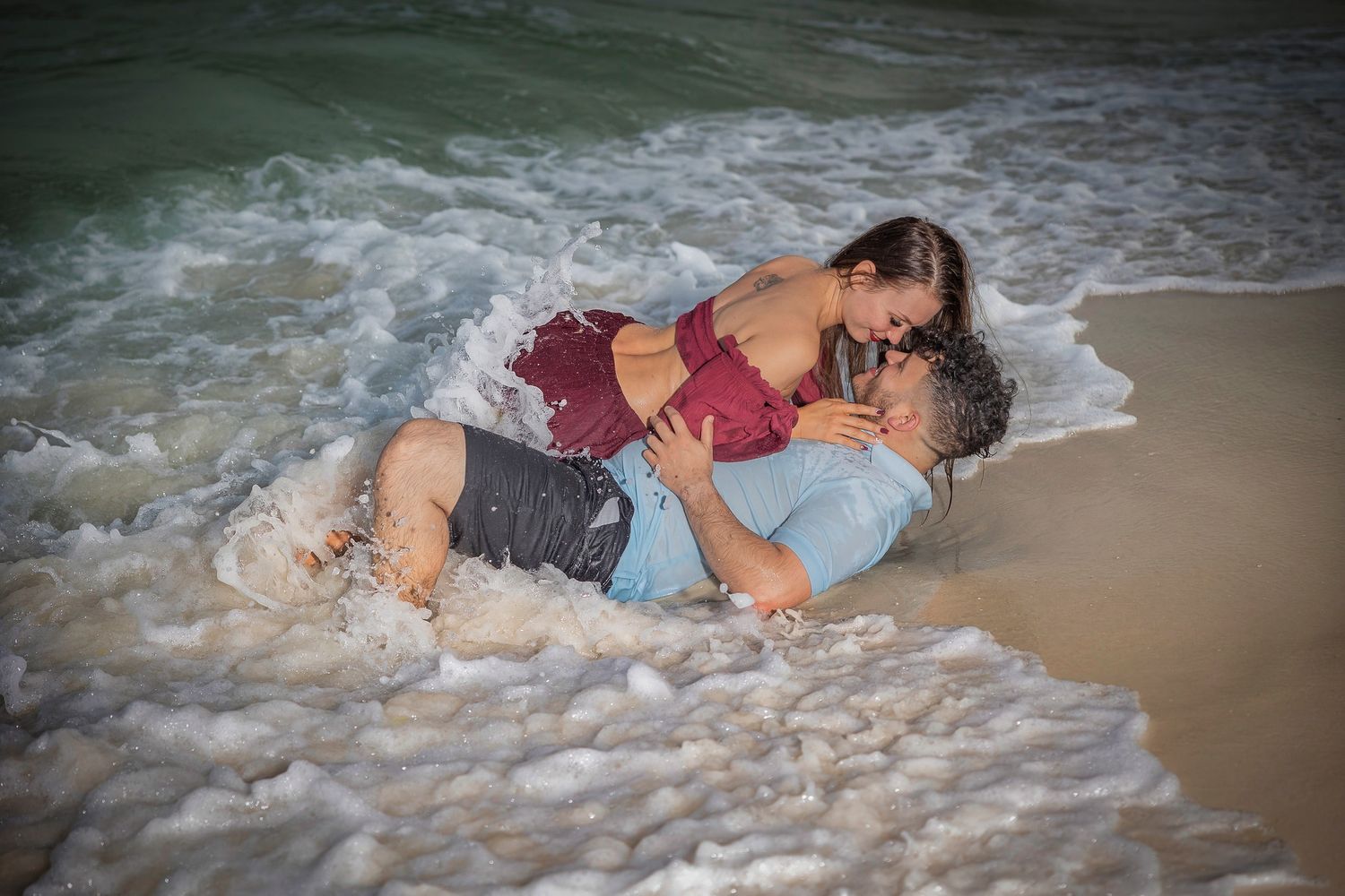 A couple laying on a sandy beach as gentle waves wash over them at sunset.