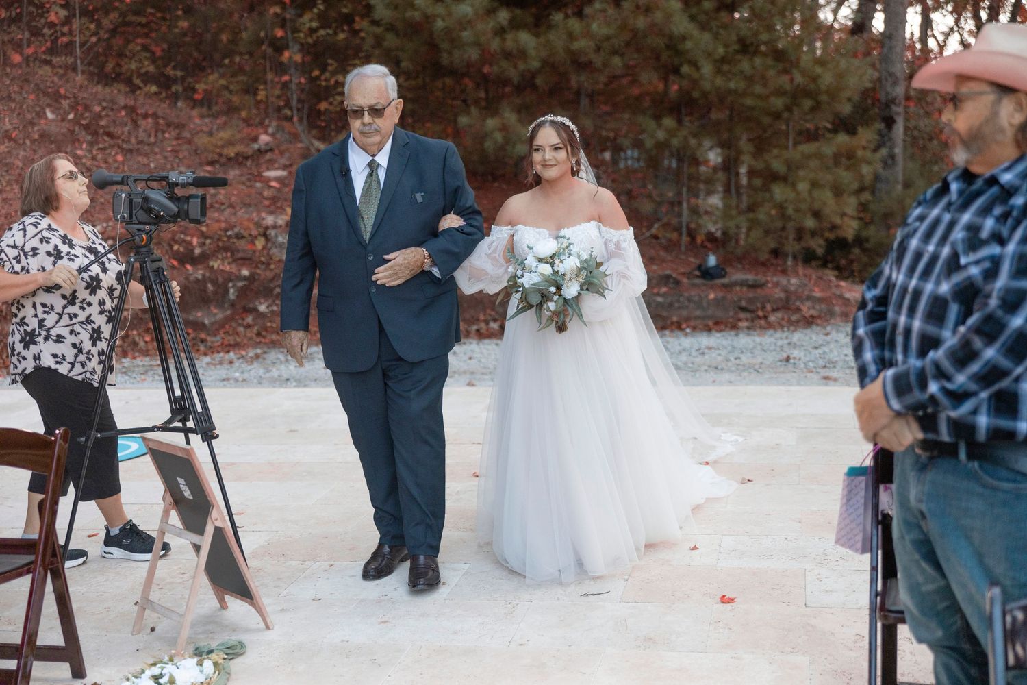 Two people walk down an outdoor aisle during a wedding ceremony as videographers capture the moment.