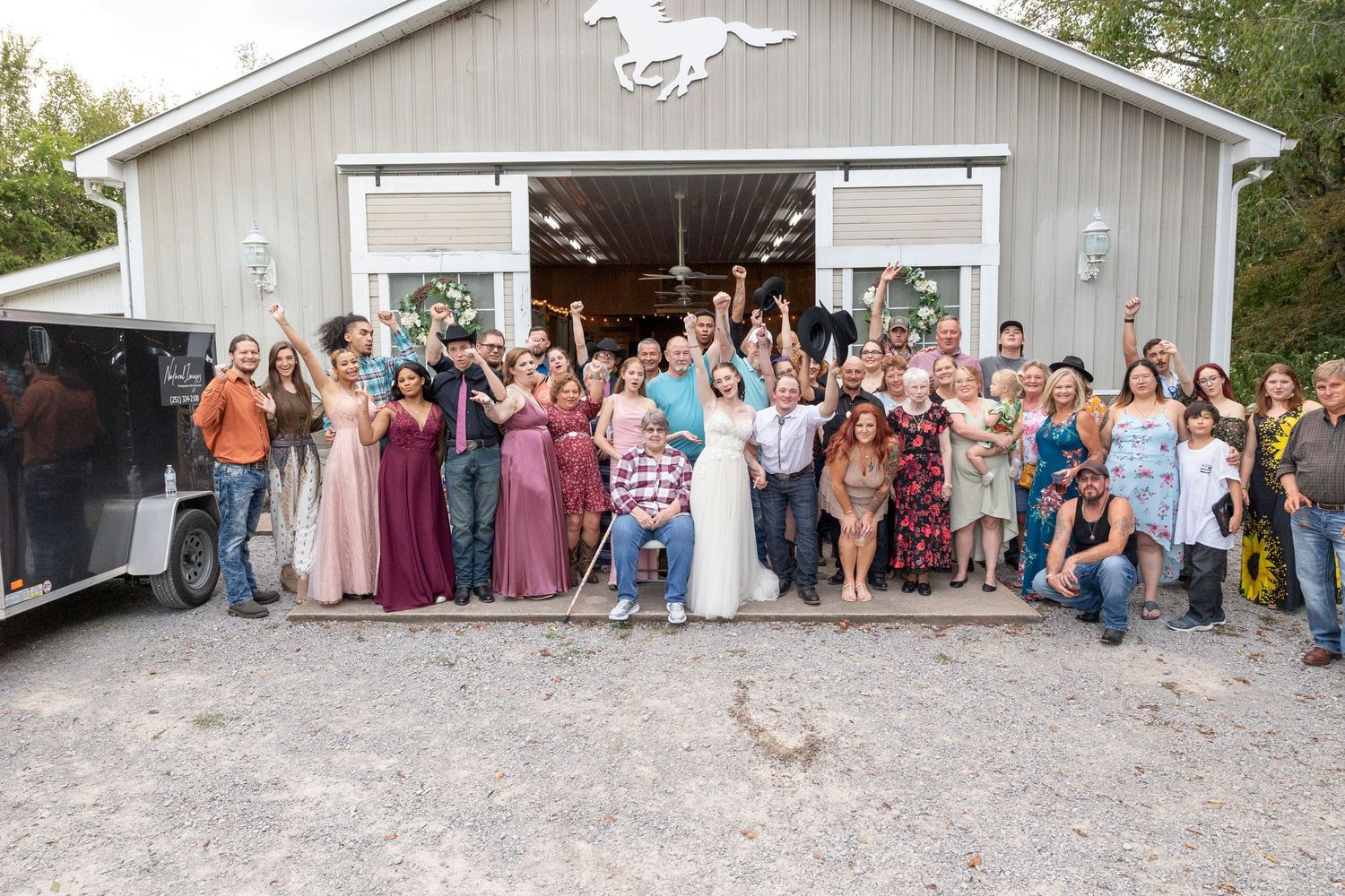 Large wedding party poses together outside a rustic barn with horse motif.