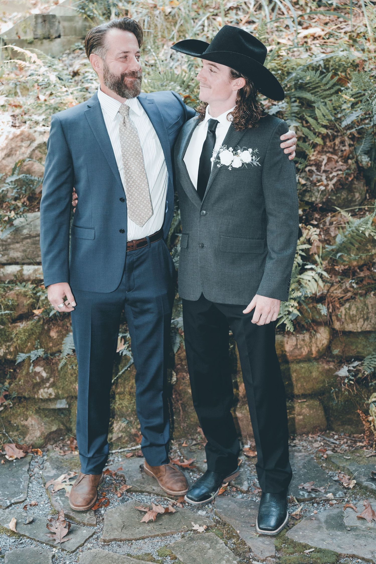 Two people in formal suits share a happy moment against a stone wall with greenery at what appears to be a wedding.
