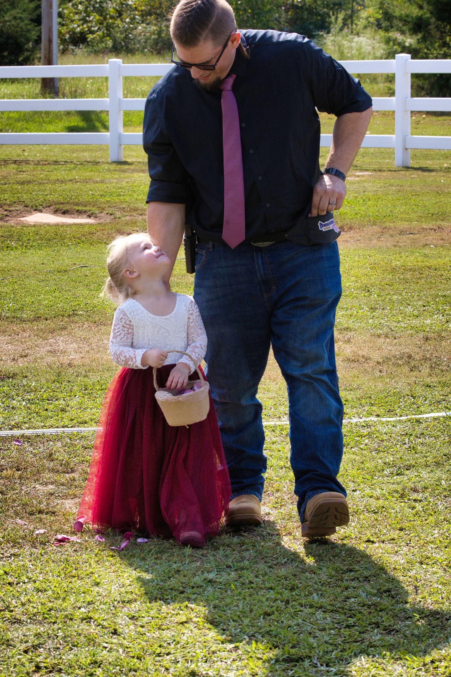 A person in a black shirt and pink tie stands with a small child in a red tulle skirt in front of a white fence.