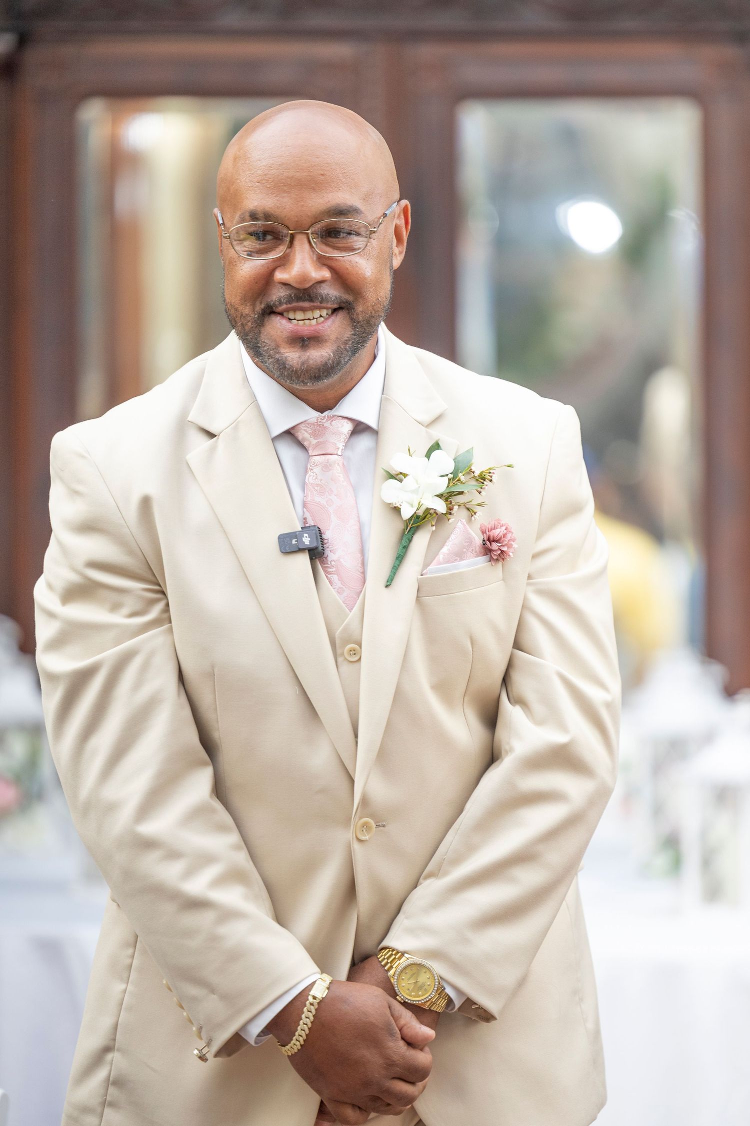 Wedding guest in cream colored suit with pale pink tie and white boutonniere smiles warmly at special occasion.