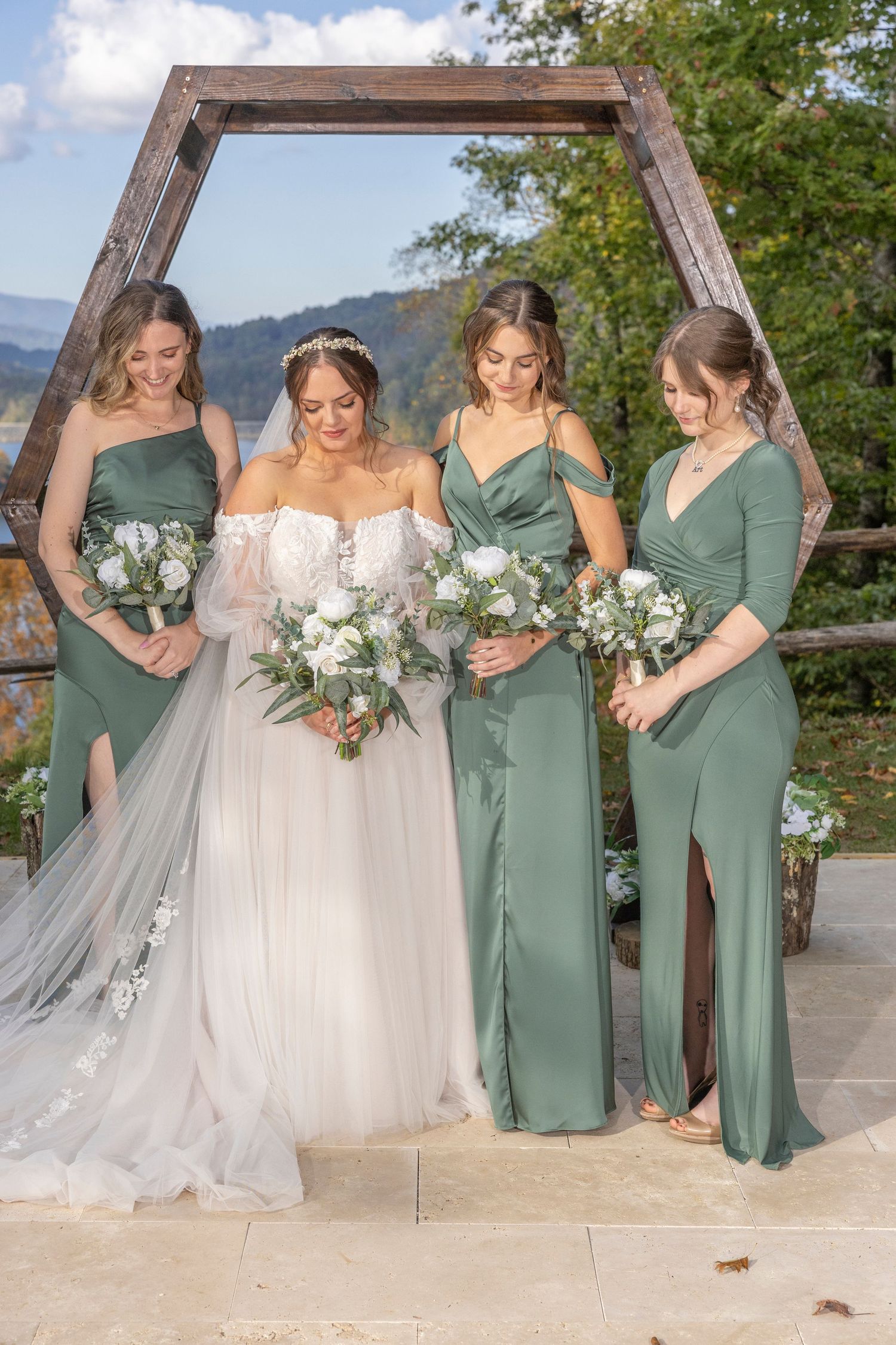 Beautiful bridal party in sage green dresses poses under wooden hexagon arch against mountain landscape backdrop.