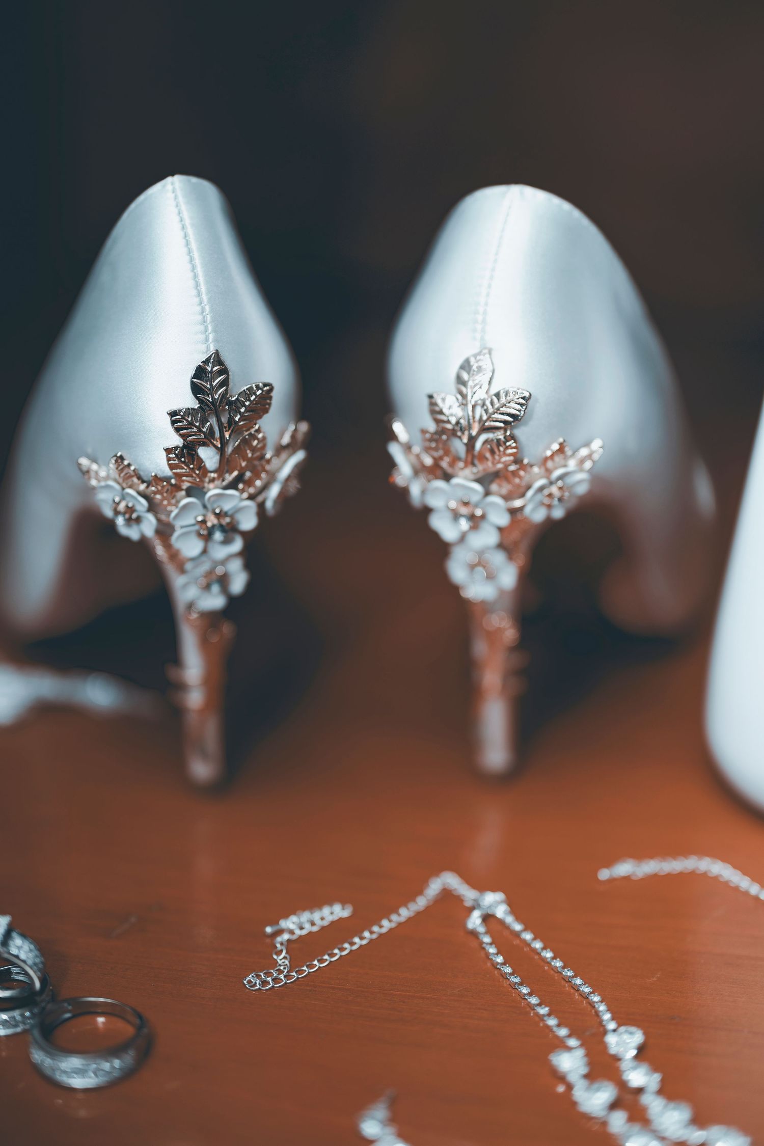 Elegant silver wedding heels with decorative crystal and leaf embellishments on display with matching jewelry on wooden surface.