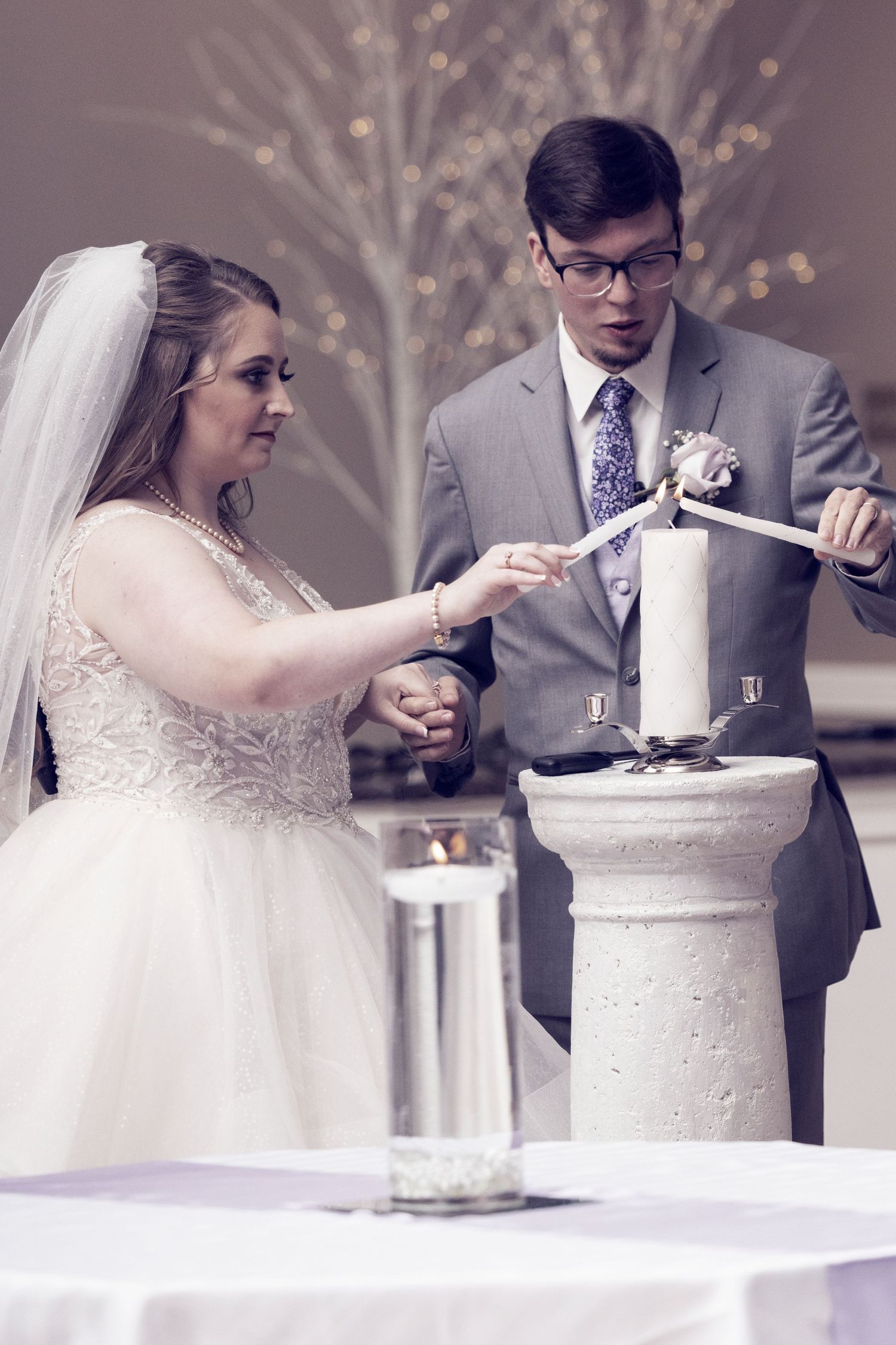 Couple performs unity candle lighting ceremony at wedding against a backdrop of twinkling lights.