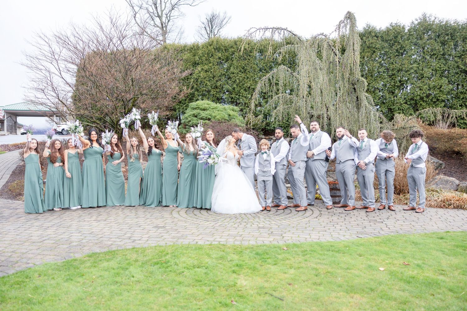 Wedding party in seafoam green dresses and gray suits poses outdoors with newlyweds on paved garden path.