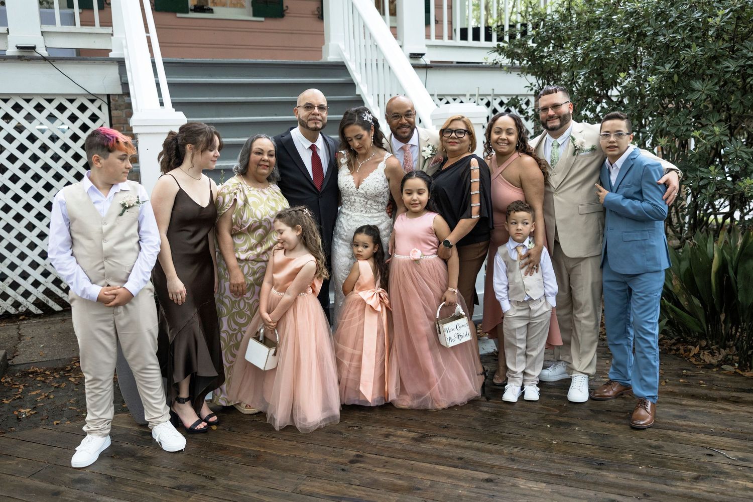A wedding party wearing coordinated pink, beige and blue attire poses together on a wooden deck patio.