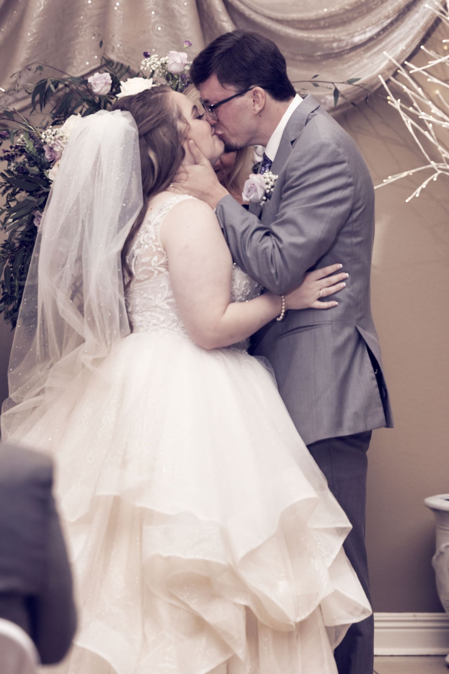 A couple shares a romantic kiss during their wedding ceremony under elegant draped fabric and floral decor.