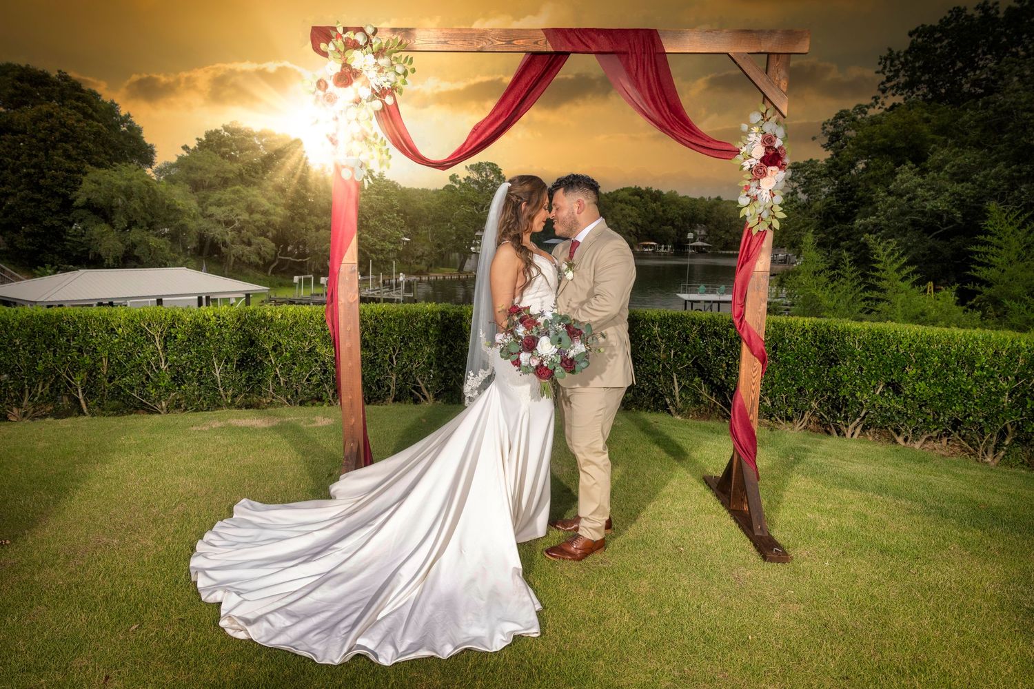 A wedding couple shares a romantic moment under a red-draped wooden arch at sunset in a lush garden setting.