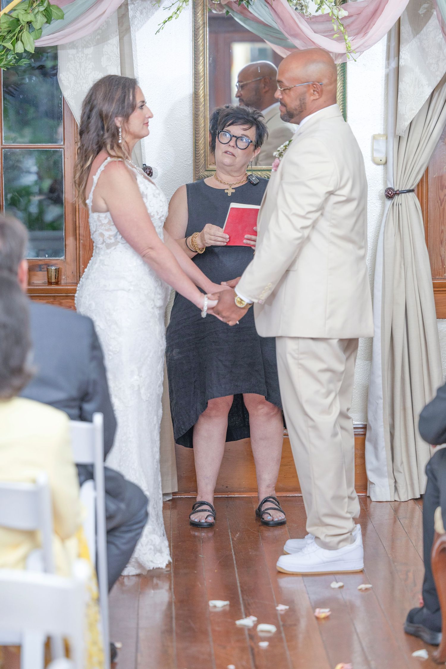 A couple holds hands during their wedding ceremony with an officiant in black standing between them.