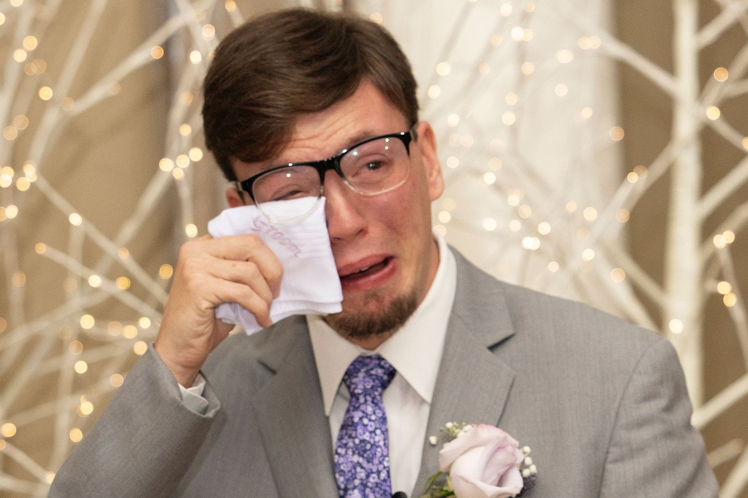 An emotional wedding moment captured as someone wearing a grey suit and purple tie dabs tears with a tissue.