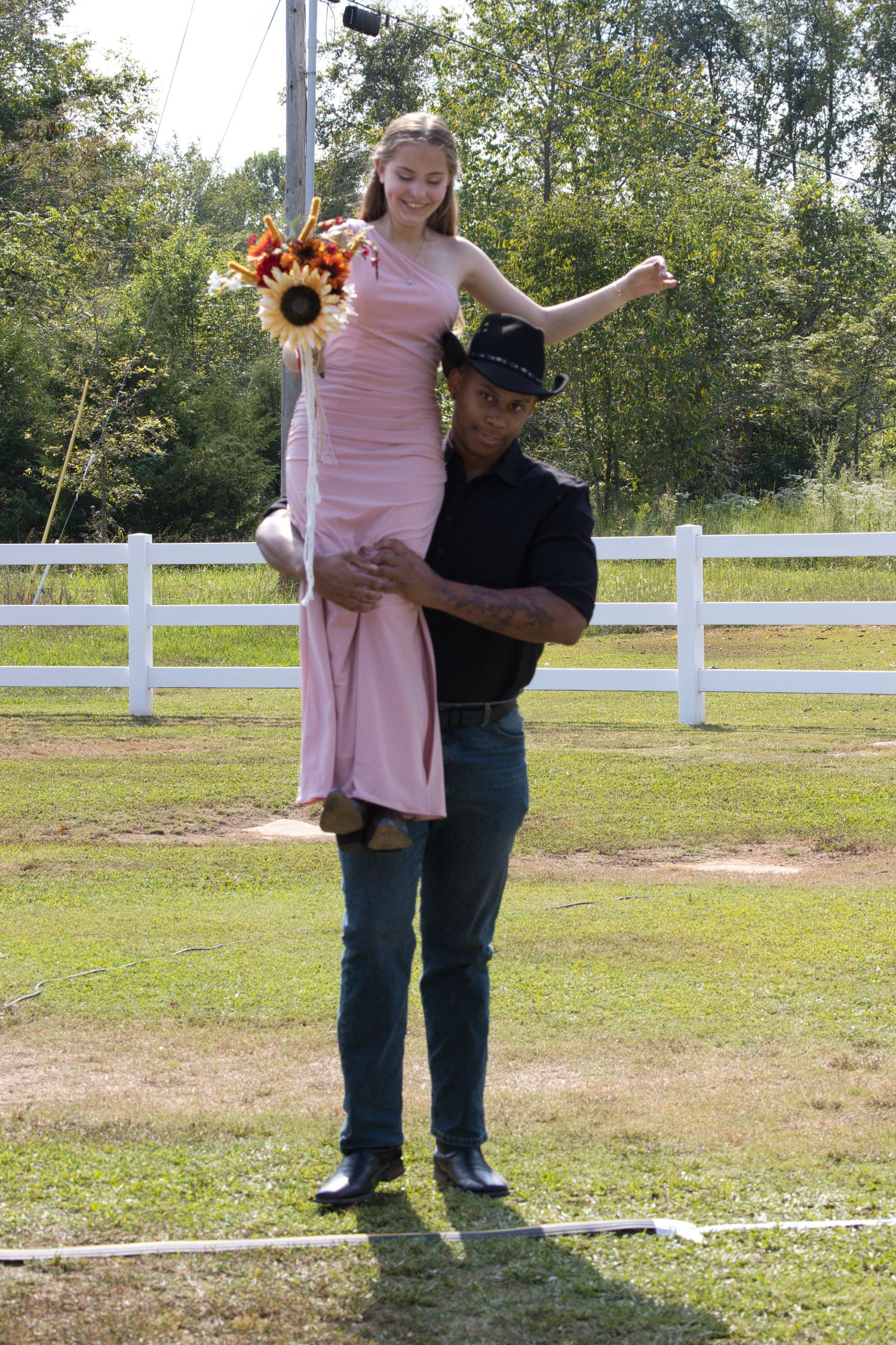 A couple poses playfully in front of a white fence, with sunflowers in their wedding attire.