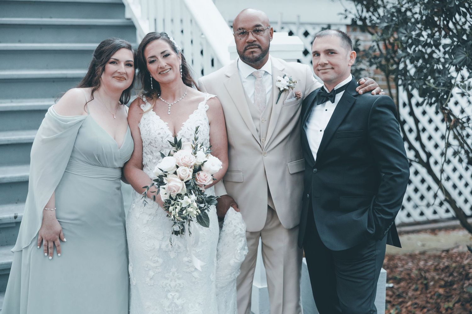 Wedding party of four people in formal attire standing together outdoors with the bride holding a white bouquet.
