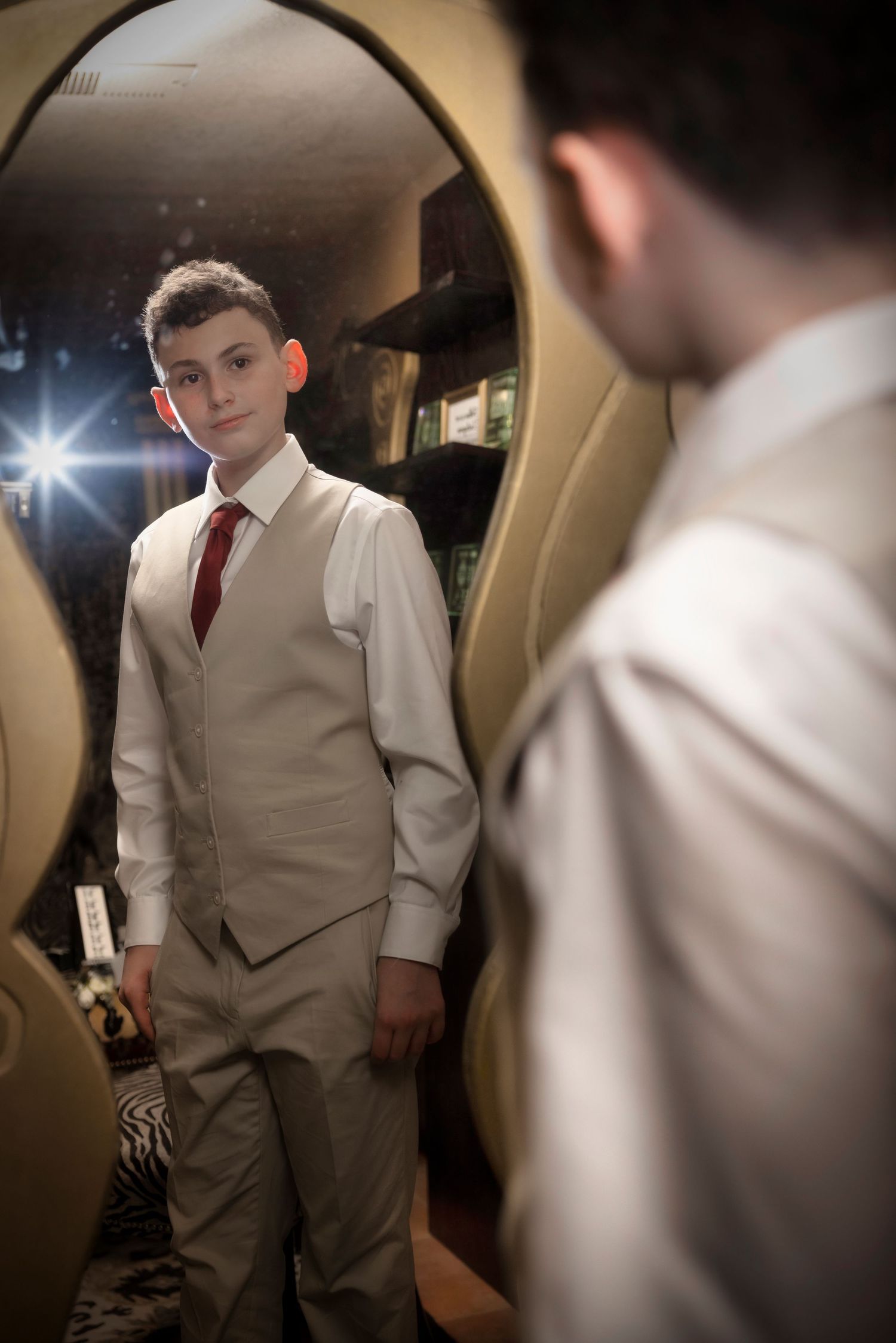 A person checks their formal attire in a mirror, wearing a tan vest and red tie with white dress shirt.
