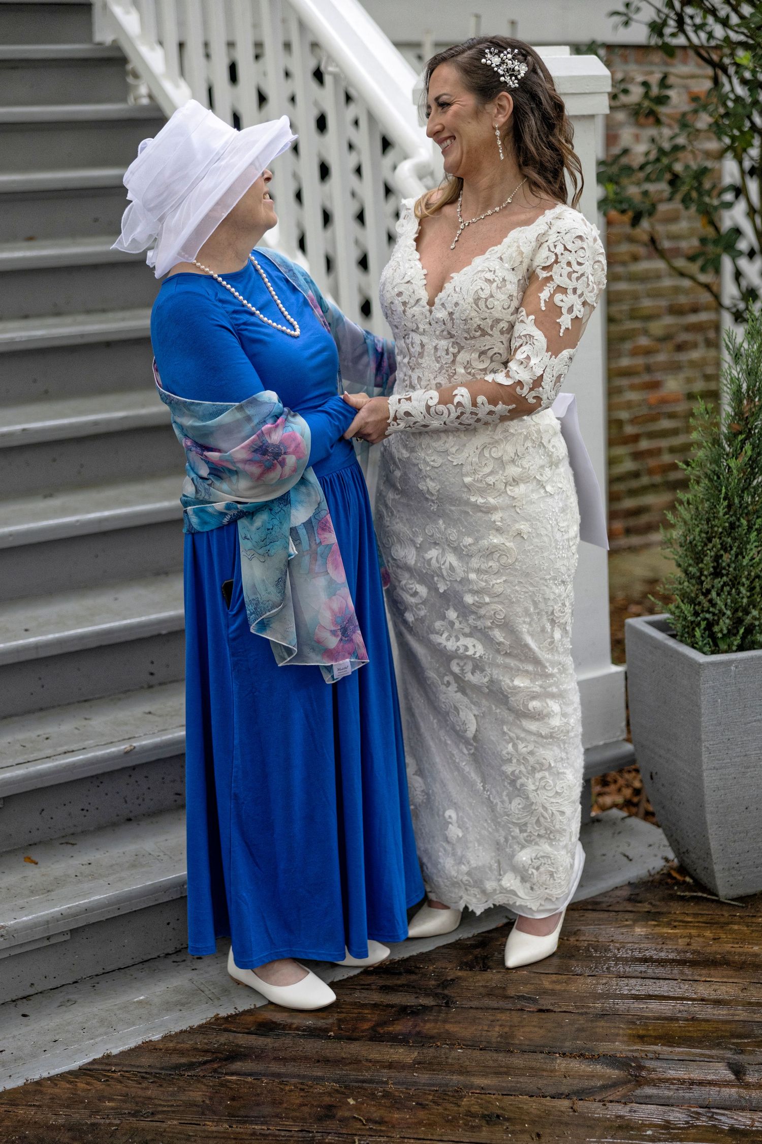 Two people in elegant attire stand on wooden steps, wearing a white lace gown and blue flowing dress with floral accents.