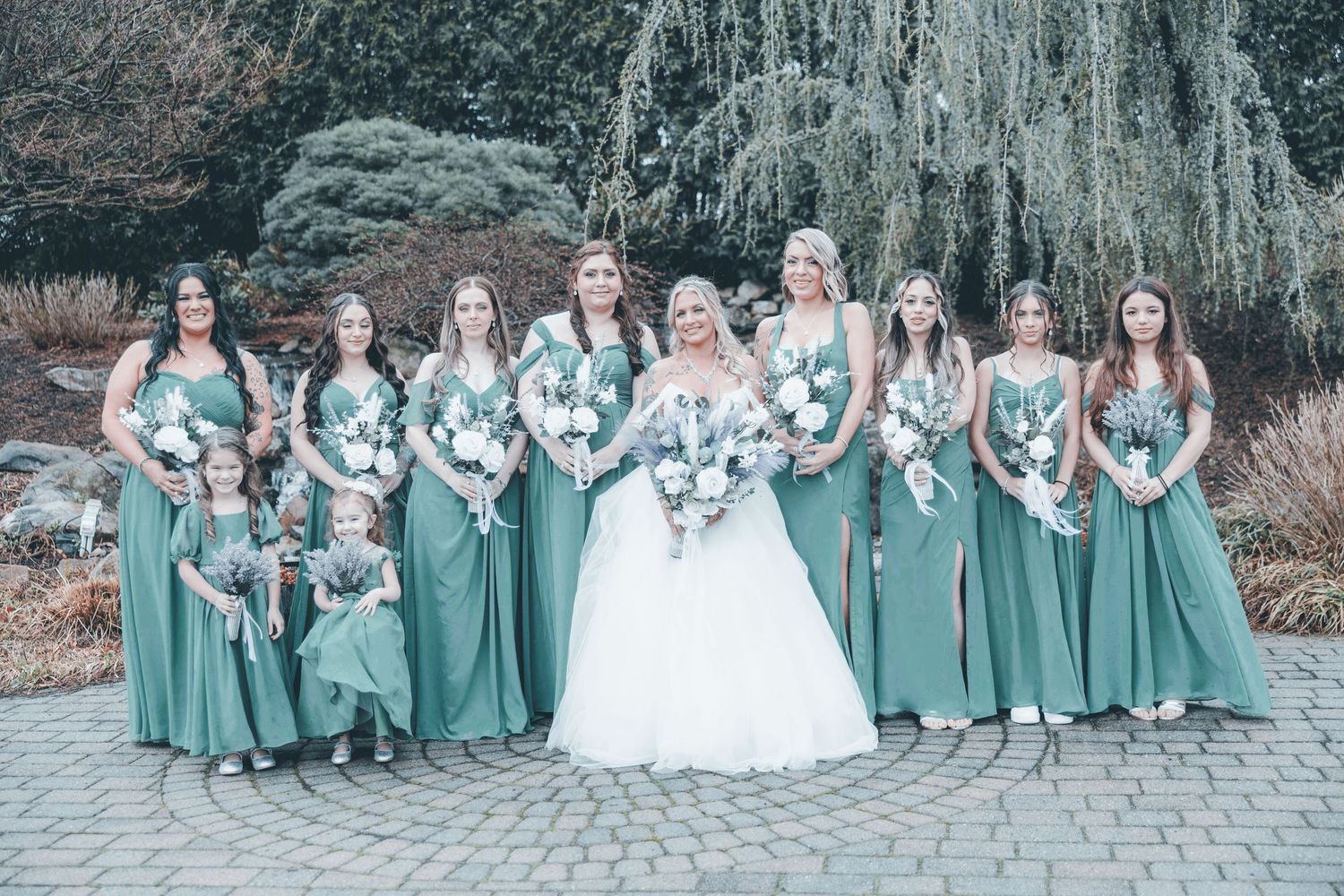 A wedding party poses outdoors with bridesmaids in sage green dresses and white bouquets on a stone walkway.