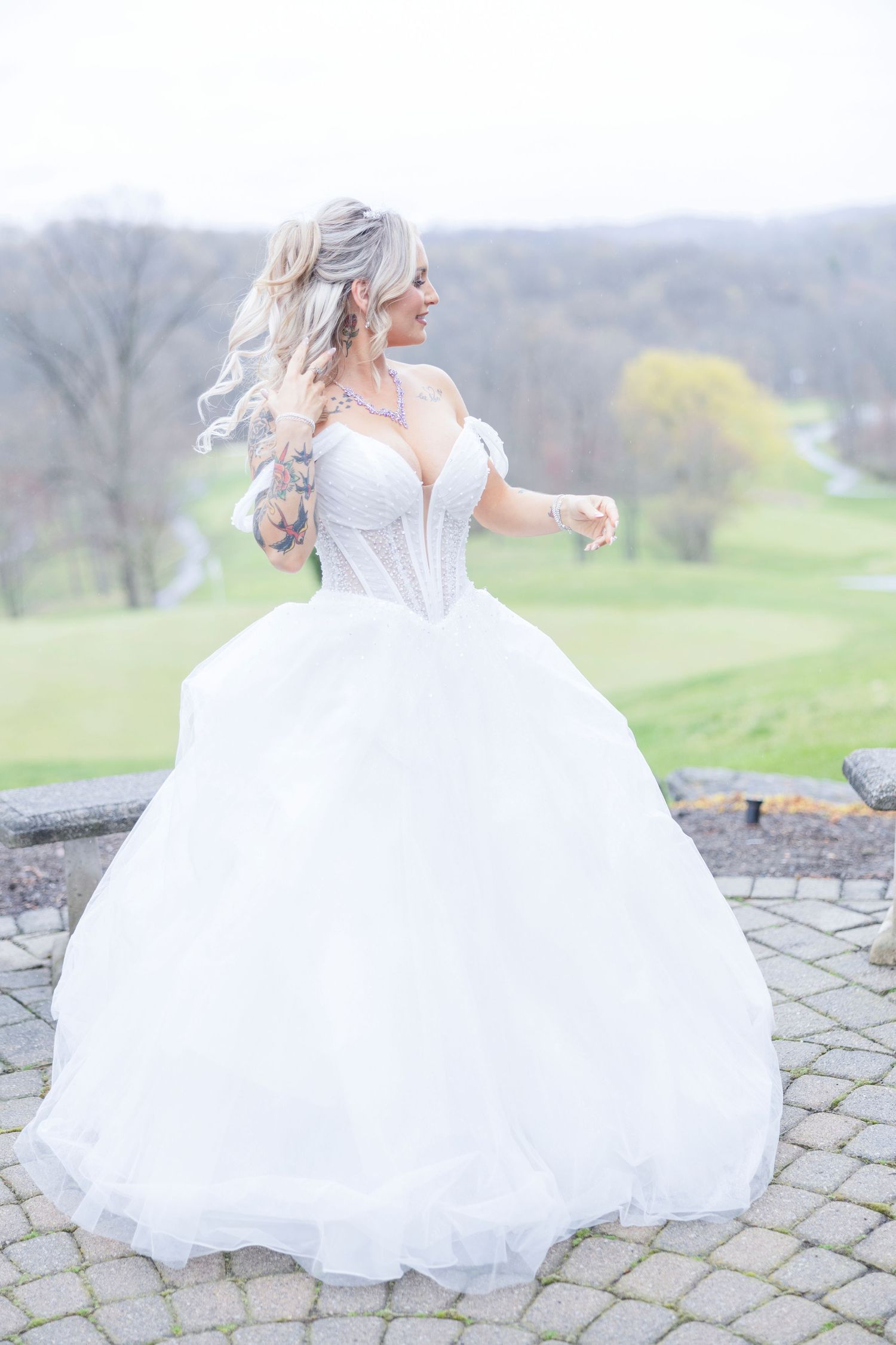 A bride in a flowing white ballgown and corset bodice poses gracefully on a stone patio overlooking rolling hills.
