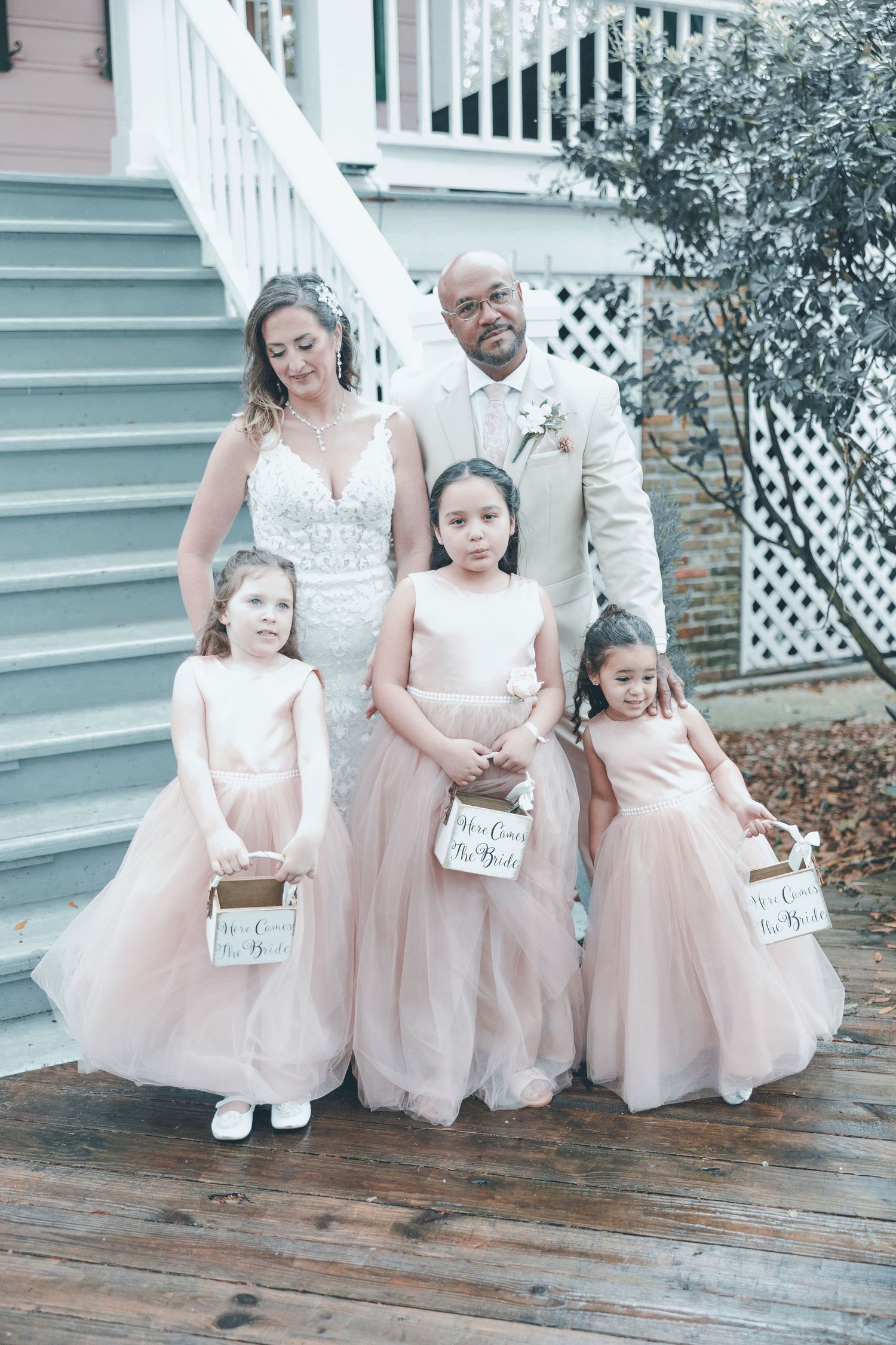 A wedding party in formal attire stands on wooden stairs, with the bride in white and three flower girls in pink dresses.