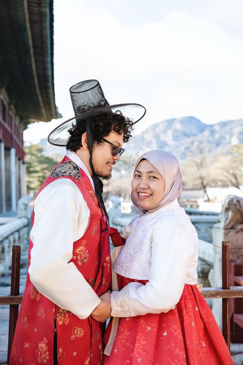 A couple in traditional red and white Korean hanbok outfits share a sweet moment together outdoors.