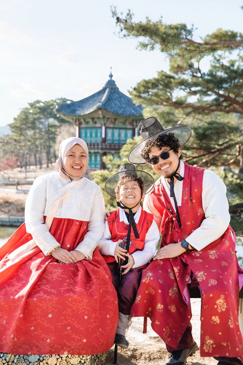 Three people in traditional red and white Korean hanbok outfits smile and laugh together in a sunny outdoor park setting.