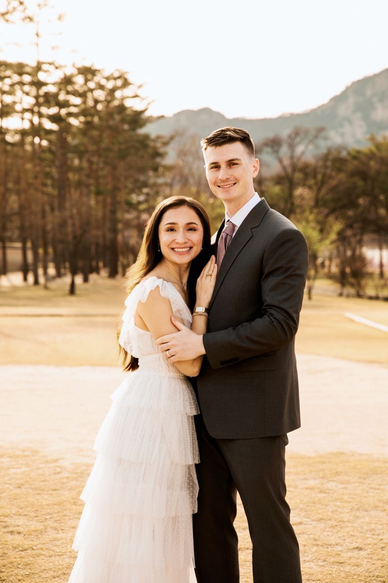 A couple shares an intimate moment in a golden-lit field with mountains and pine trees in the background.
