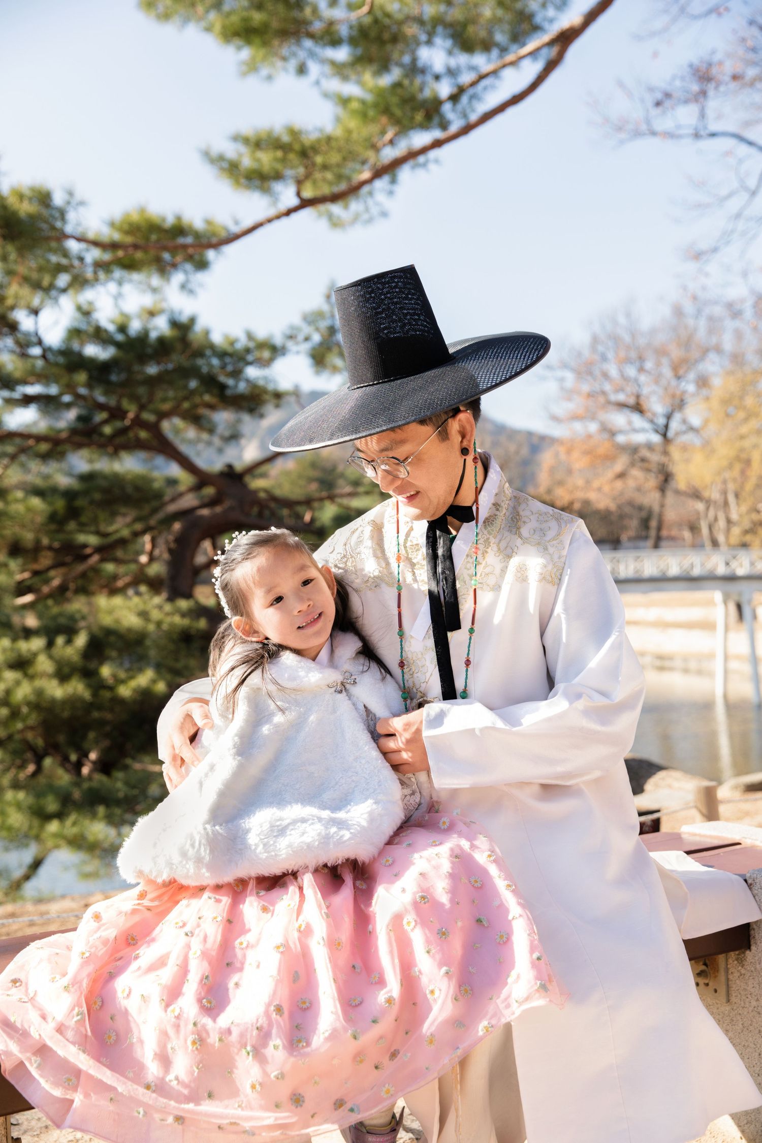People wearing traditional Korean hanbok clothing embracing in front of a scenic lake and temple.